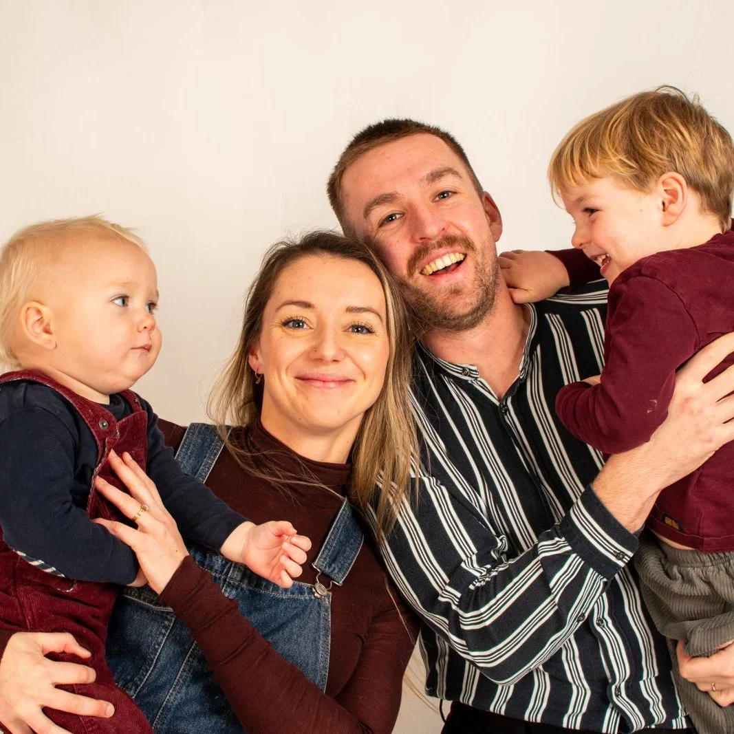 Family of four, two children, and two adults smiling together, sitting against a plain white background.