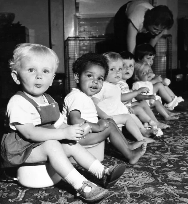 Six toddlers sitting on toilets in a row with a woman tending to them in the background.