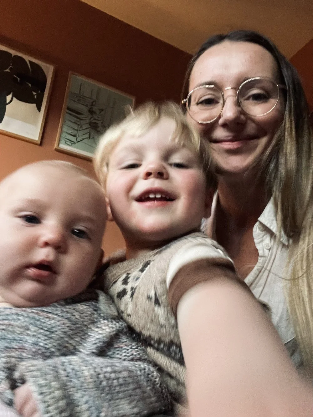 A woman with glasses taking a selfie with two young children, one with red hair and a broad smile, and the other with light hair and a neutral expression, indoors with framed artwork on the wall behind them.