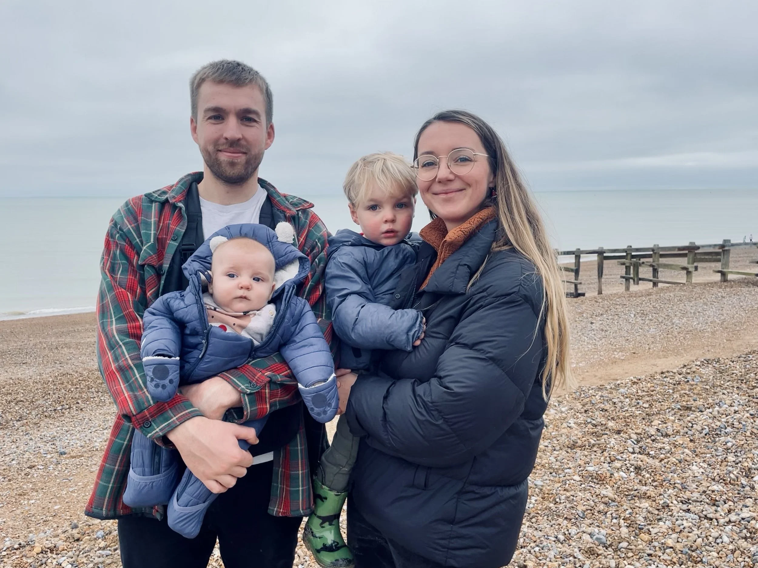 Steph Elliott—founder of Baby Pottying—and her family of four standing on a pebbled beach near the ocean, dressed in jackets, with a cloudy sky overhead.