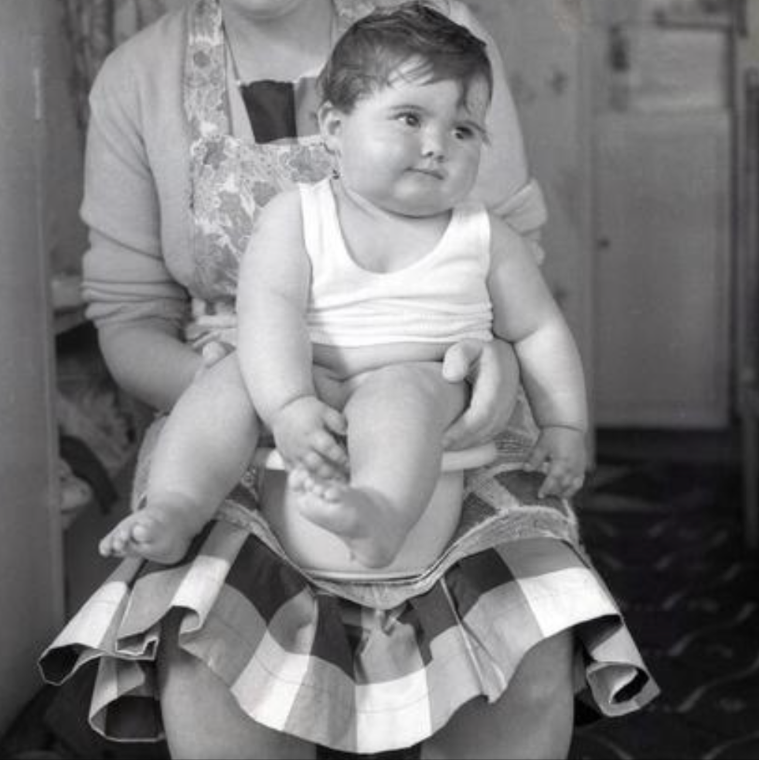Black and white photo of a young child sitting on an adult's lap on a potty, with the adult sitting behind them.
