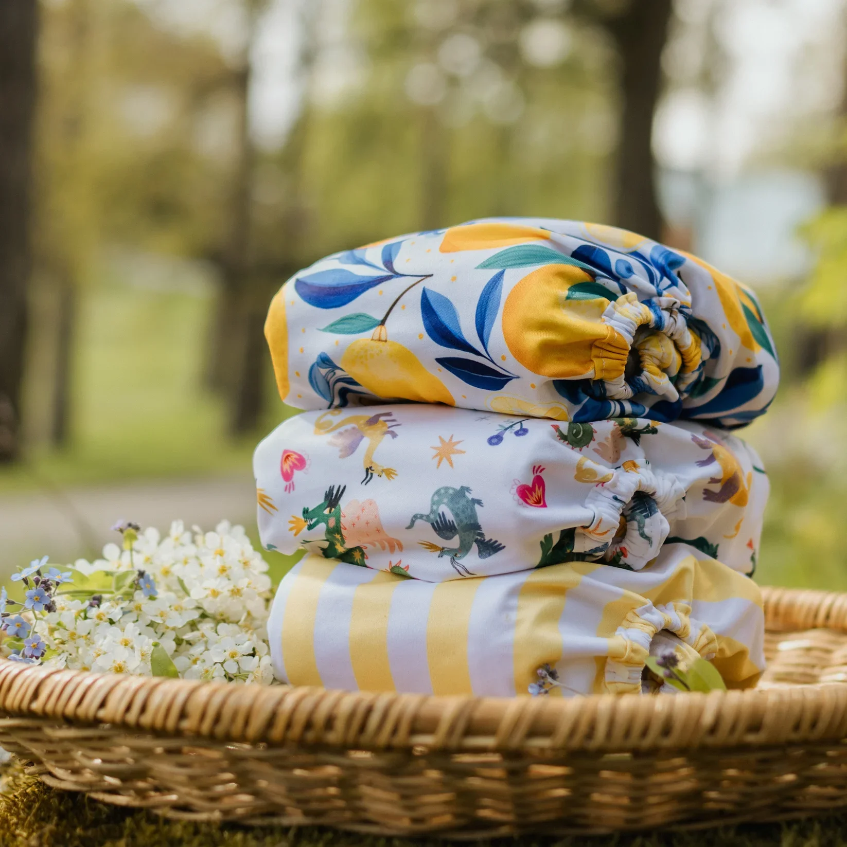 Three folded cloth diapers stacked in a woven basket outdoors with flowers nearby and trees blurred in the background.