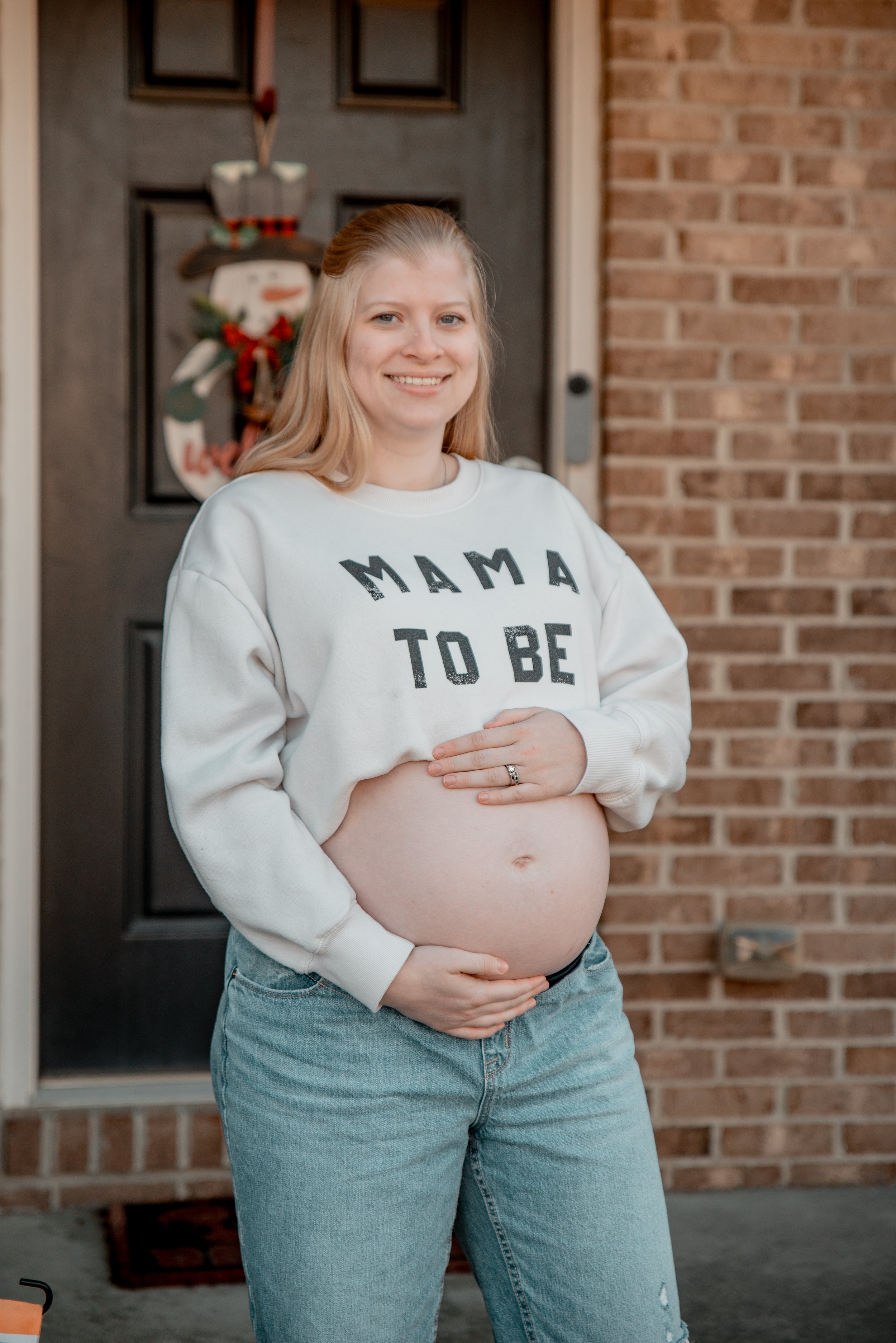 Pregnant woman standing outside in front of a brick wall and a door decorated with a holiday wreath, holding her belly with one hand and resting her other hand on top, smiling at the camera.