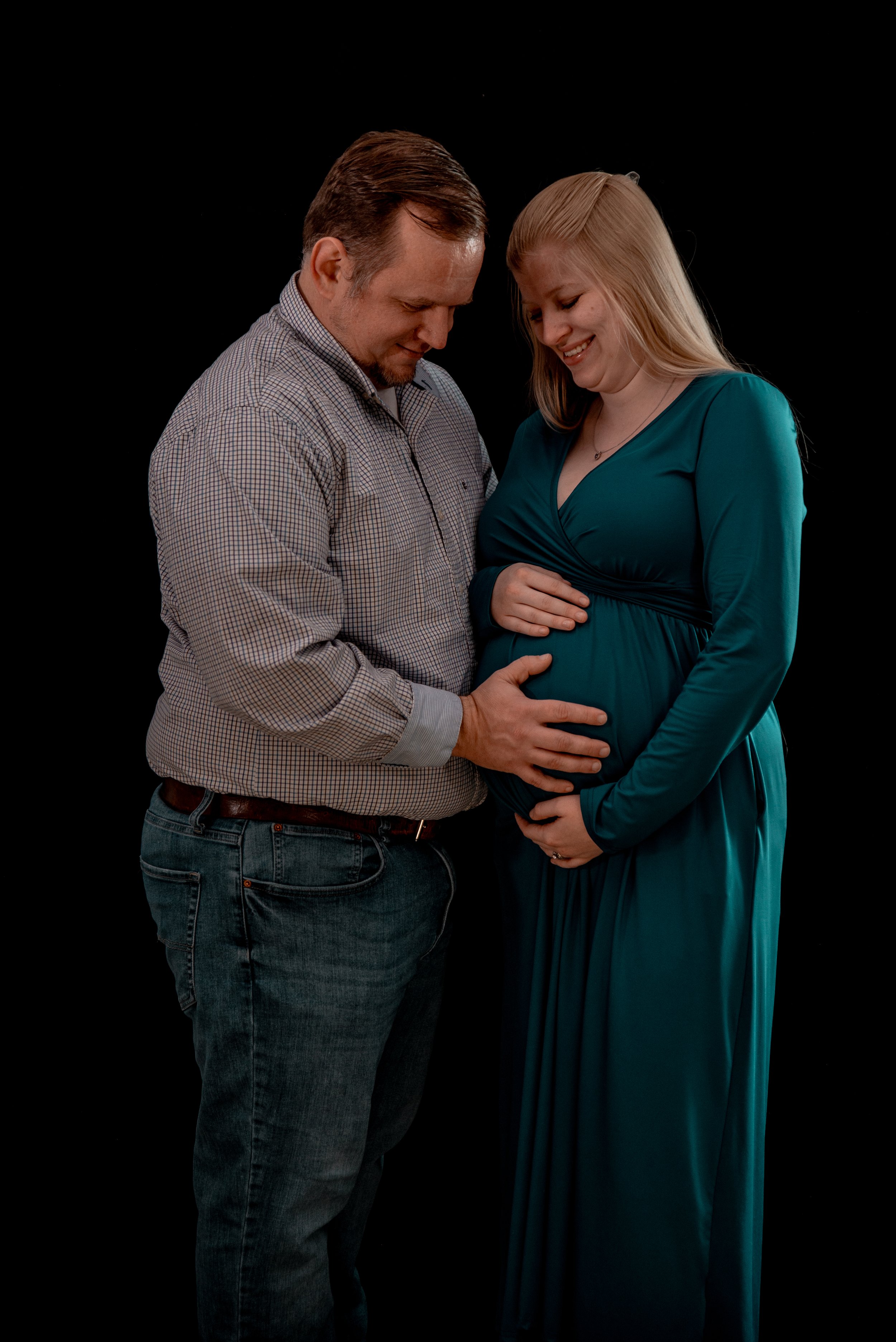 A pregnant woman and a man hold her belly and smile together against a black background.