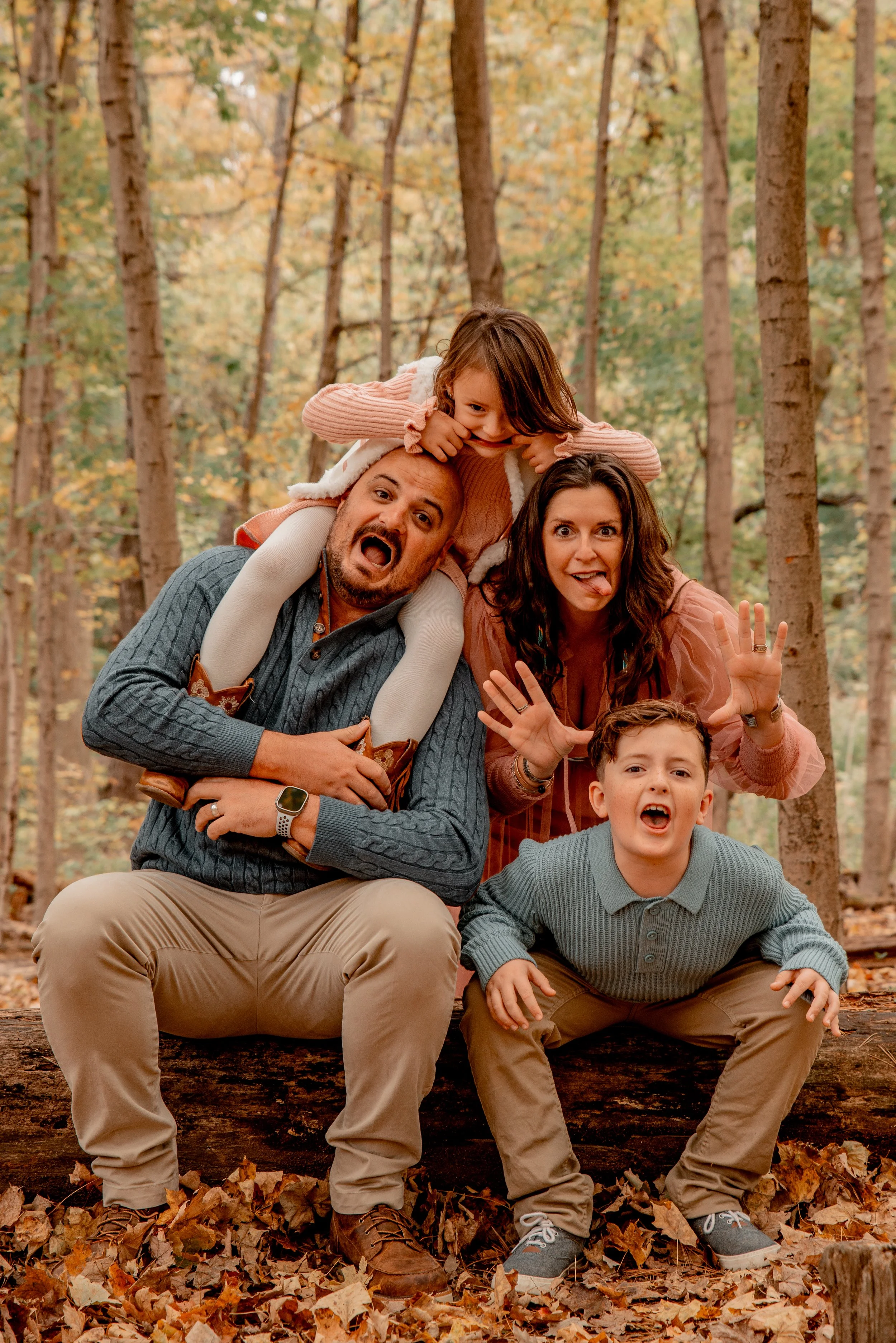 Family of four playing in a wooded forest during autumn, with fallen leaves on the ground, making silly faces and having fun.