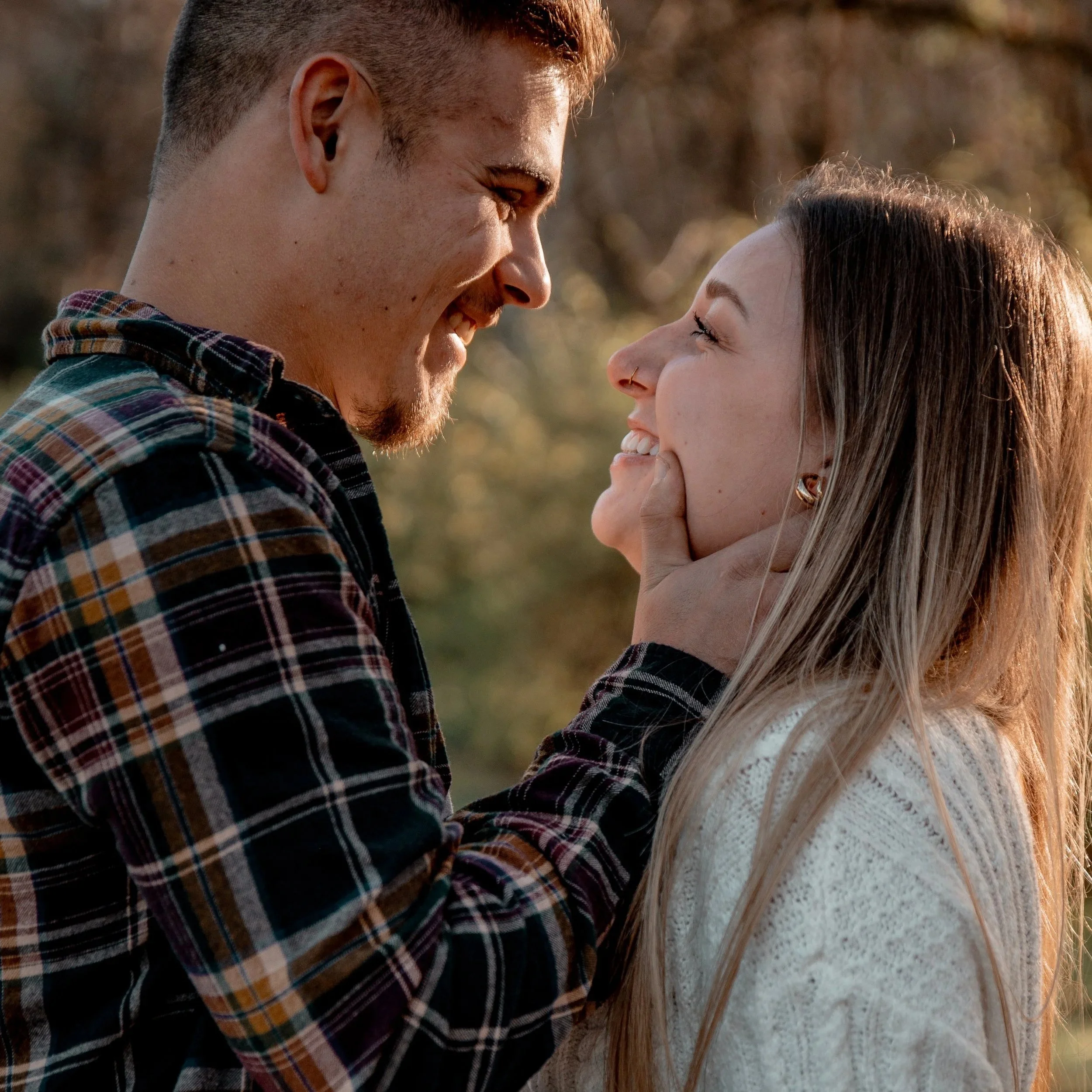 A man and a woman looking at each other, smiling, with the man gently holding the woman's chin outdoors during sunset.