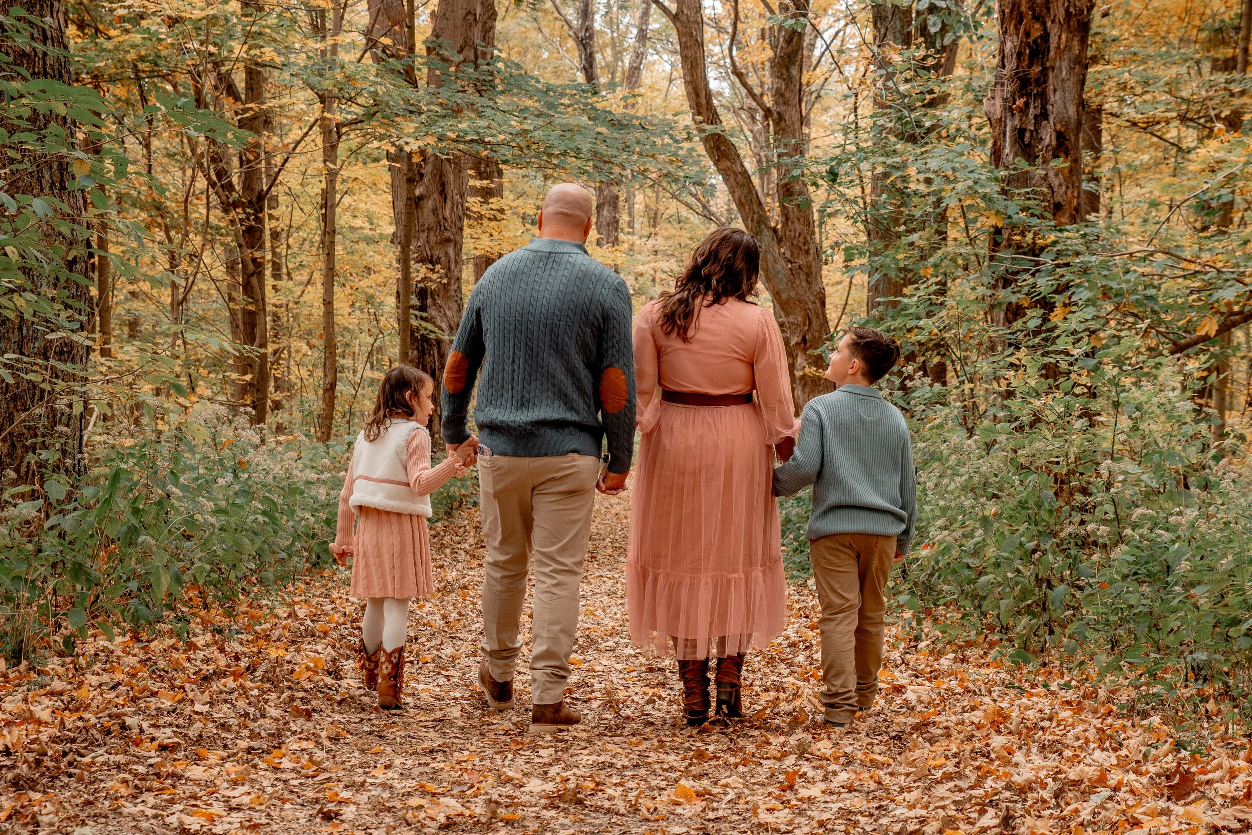 A family of four walking through a wooded area with autumn leaves on the ground. The family includes a man, a woman, and two children, a girl and a boy, holding hands and enjoying a walk in fall.