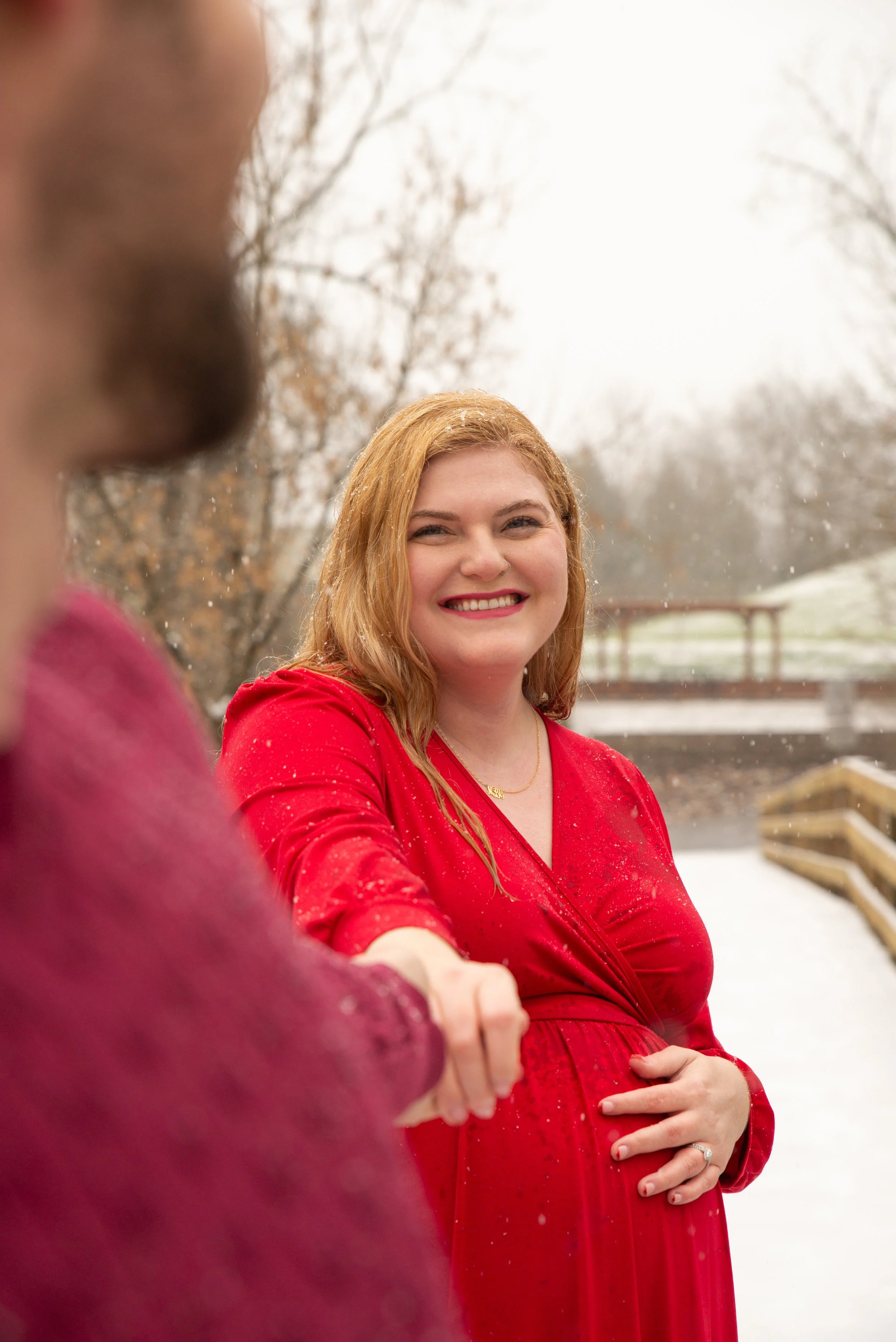 A smiling pregnant woman in a red dress walking outside during winter, holding hands with another person.