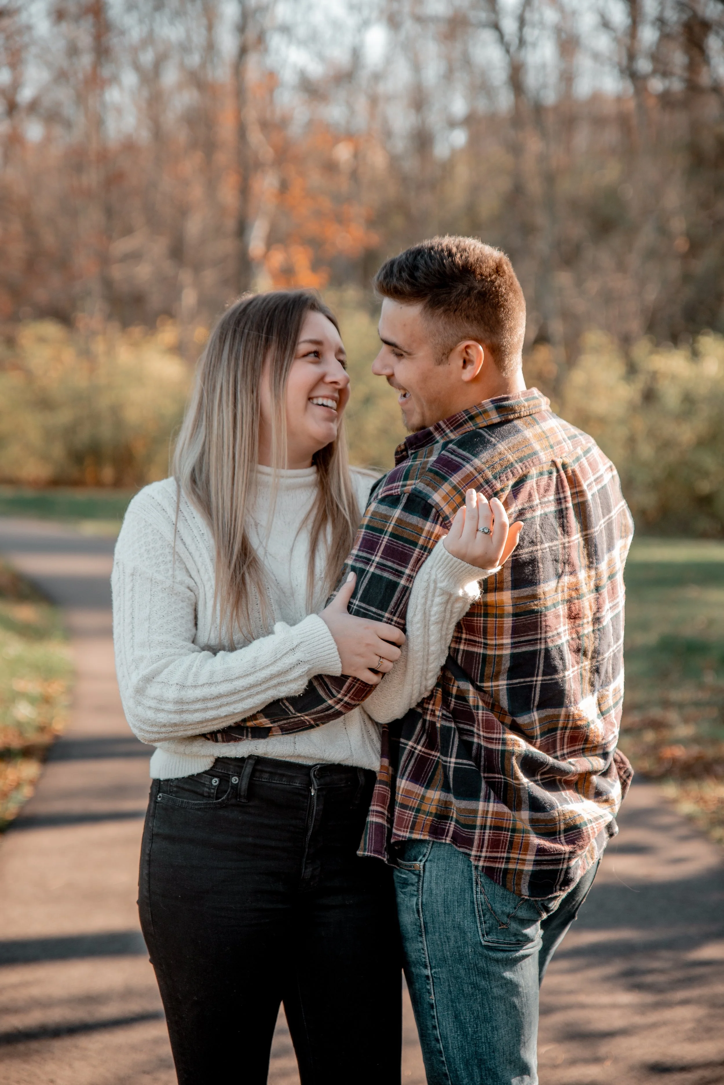 A smiling couple standing close together on a park trail during autumn, with fall foliage in the background, embracing and looking at each other.