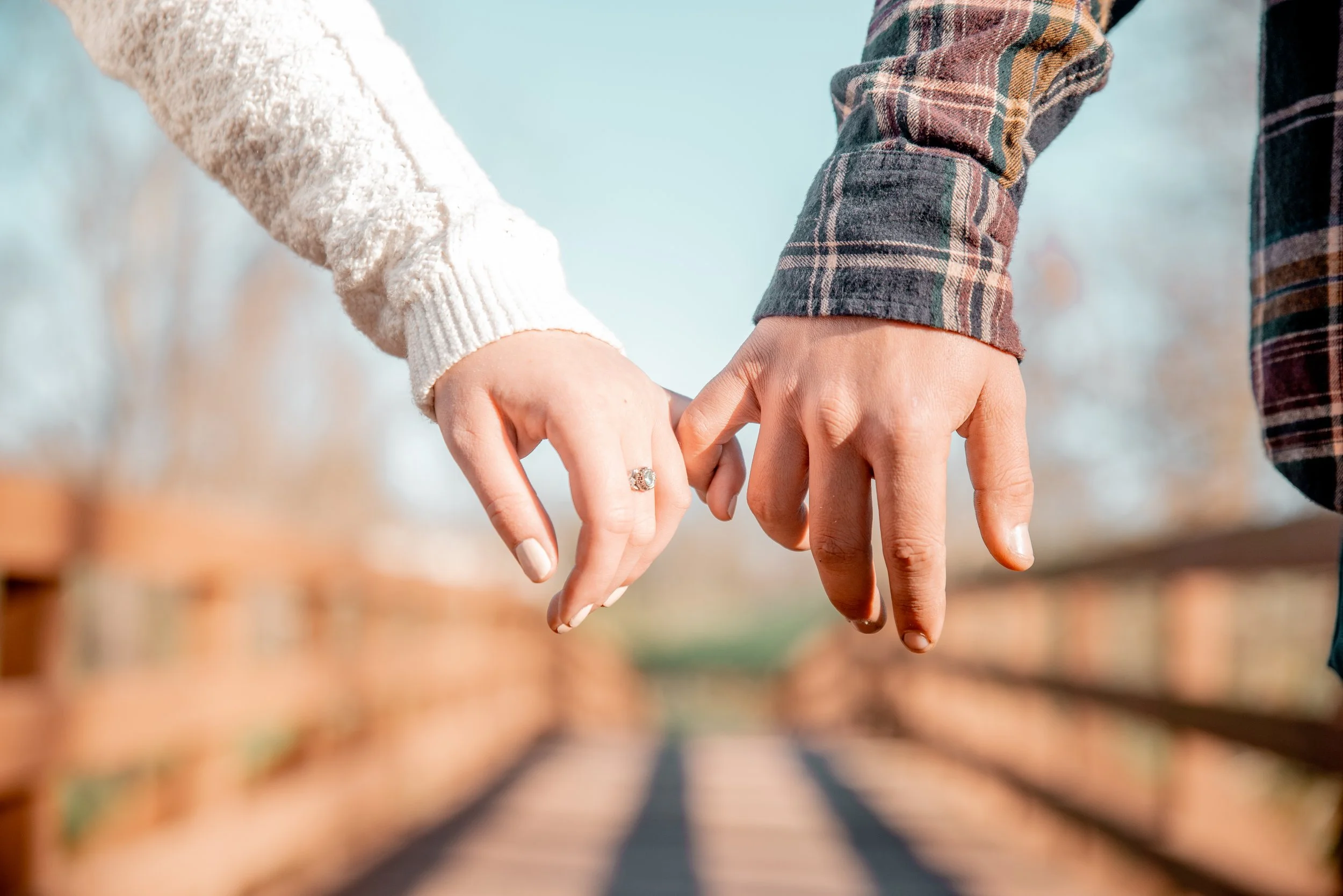 Close-up of two people holding hands on a wooden bridge outdoors, with a blurred background of trees and sky.