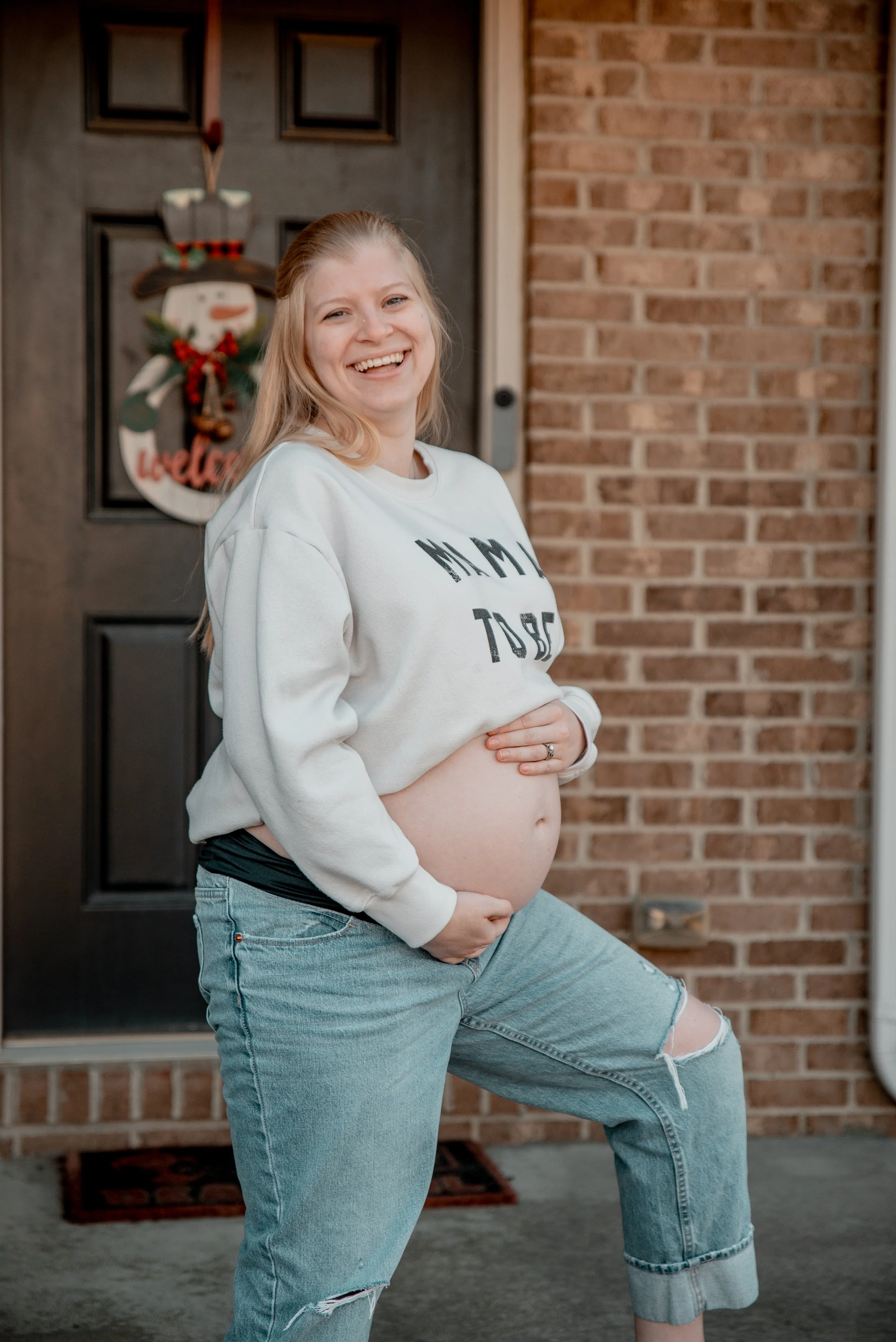 A smiling pregnant woman standing at her front door, wearing a white sweatshirt and ripped jeans.