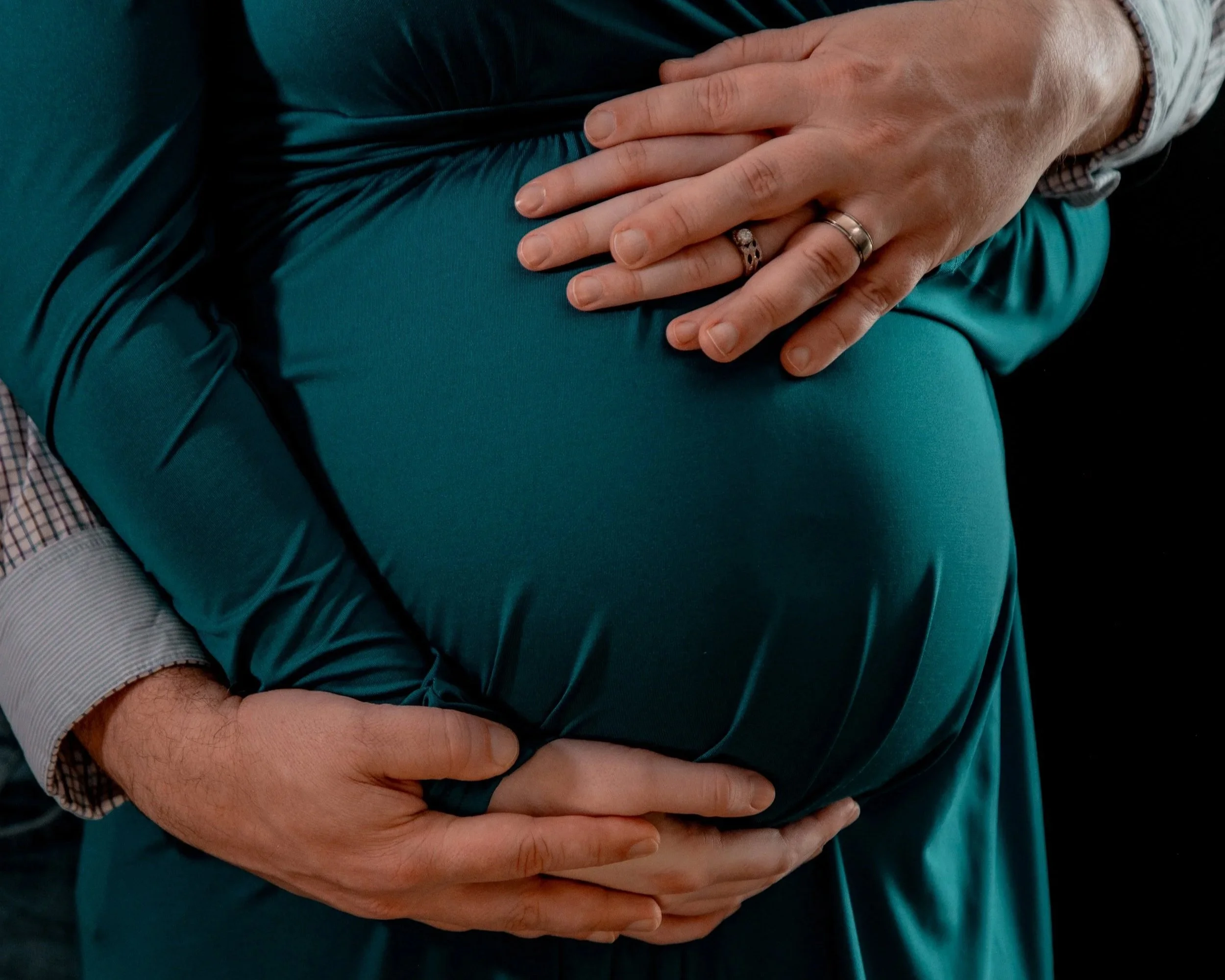 Two people holding a pregnant belly. One with hands on top, the other supporting from underneath.