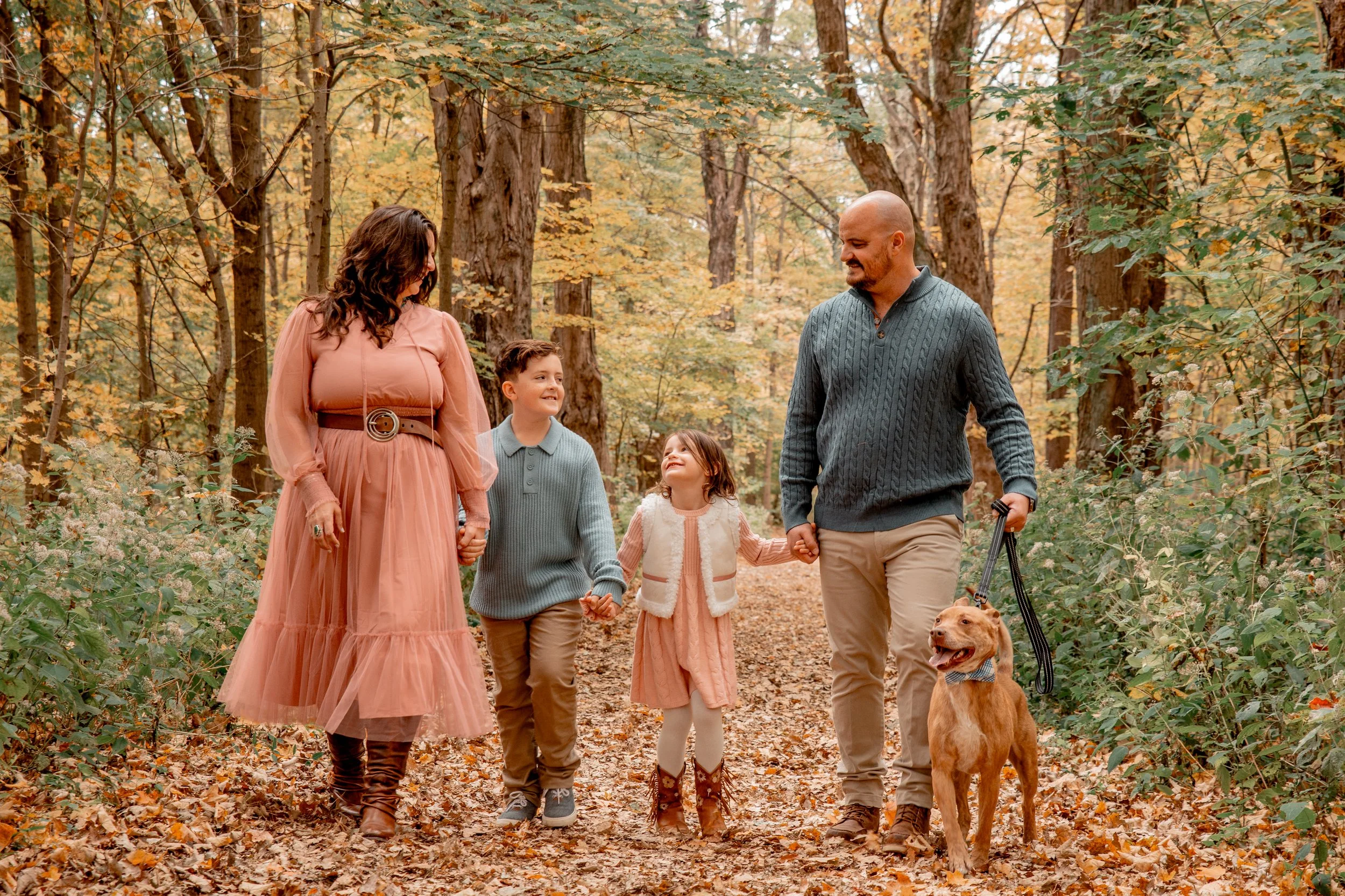 A family of five, including a woman, a man, two children, and a dog, walking hand-in-hand through a forest during fall with colorful autumn leaves on the ground and trees around them.