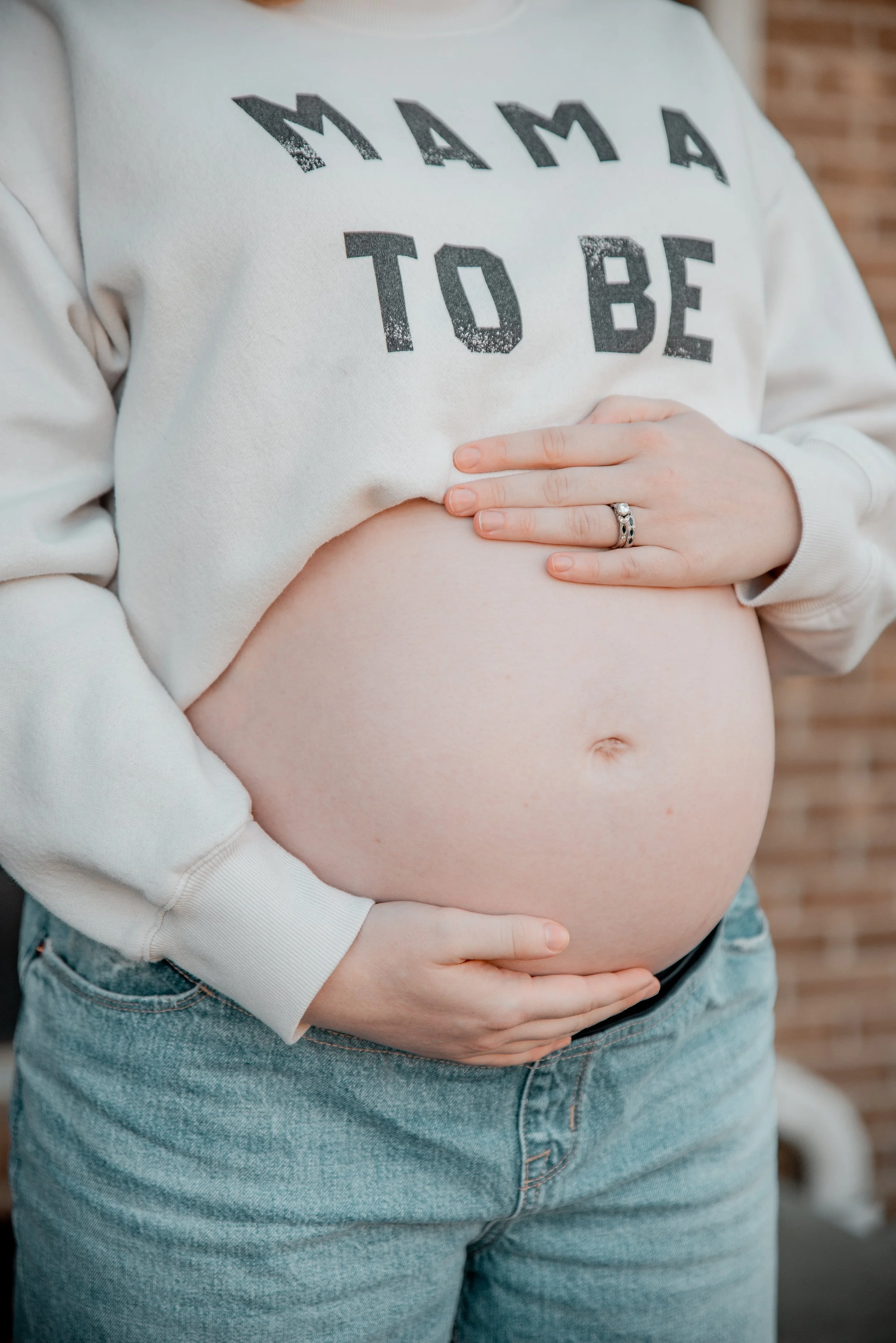 Pregnant woman holding her belly with both hands, wearing a white sweatshirt with bold text, standing against a brick wall.