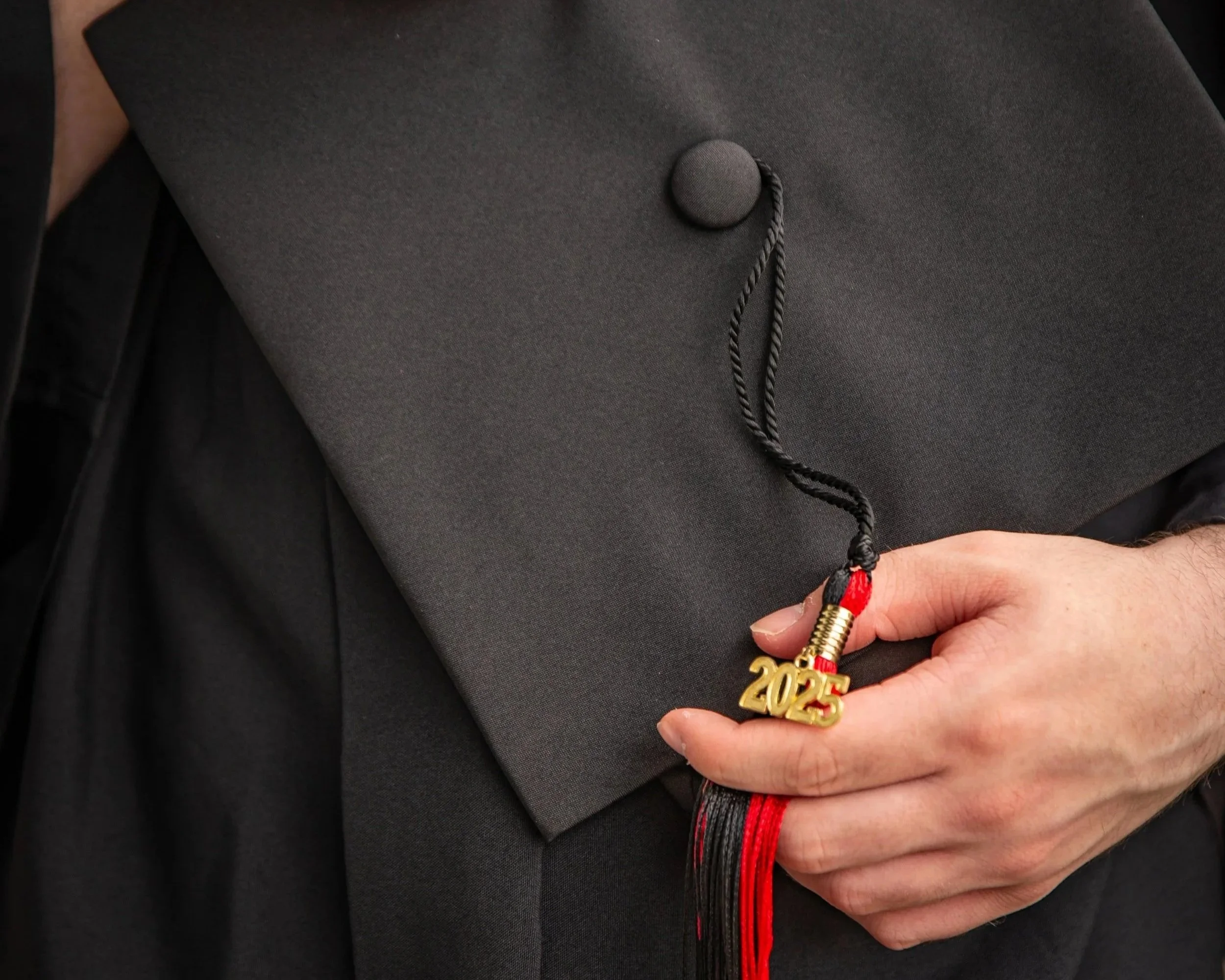 Graduation cap with a black tassel and a gold '2015' charm being held in a person's hand, dressed in a gown.