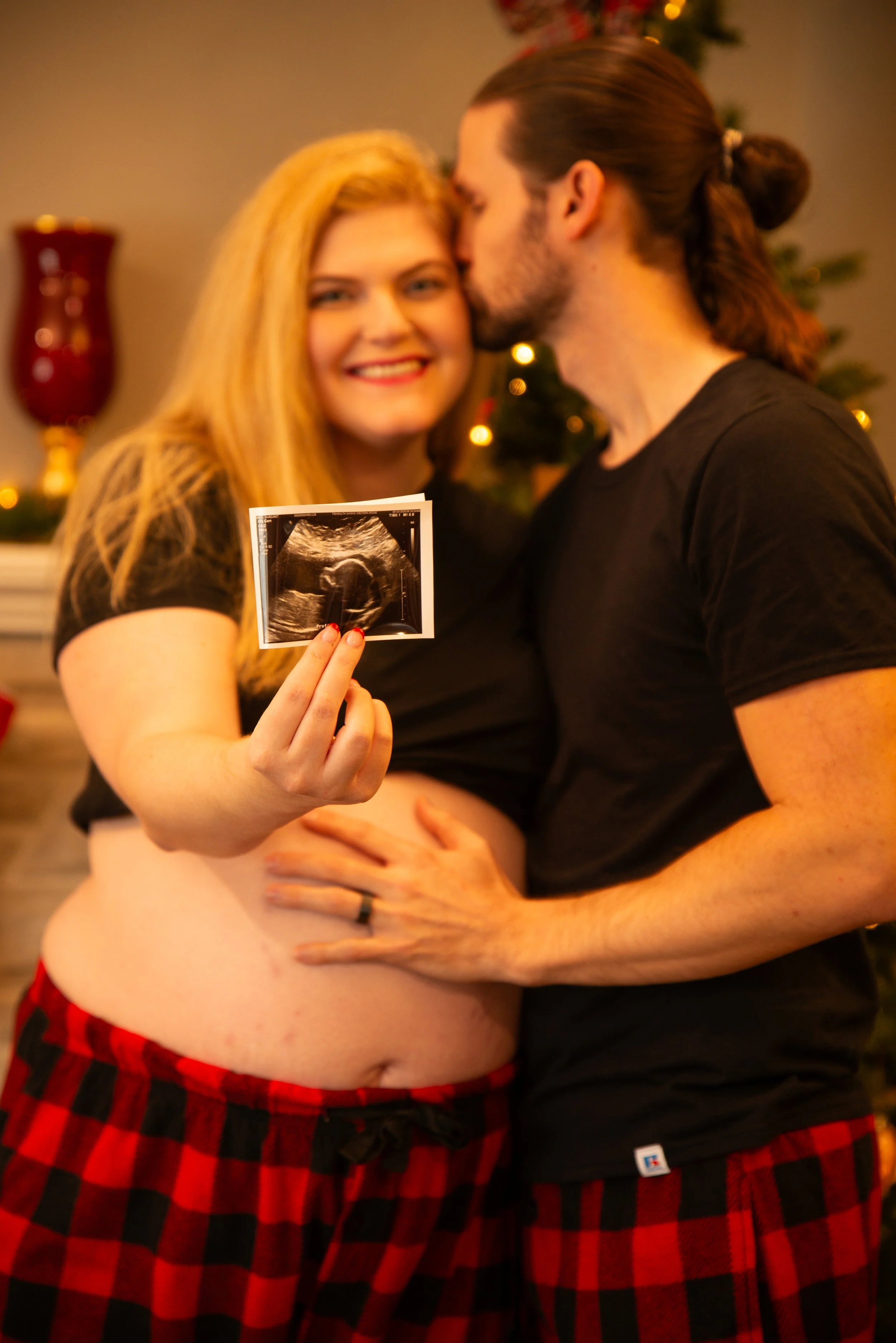 A pregnant woman holding an ultrasound picture and smiling, with a man hugging her. They are celebrating Christmas, as seen from the holiday decor in the background.