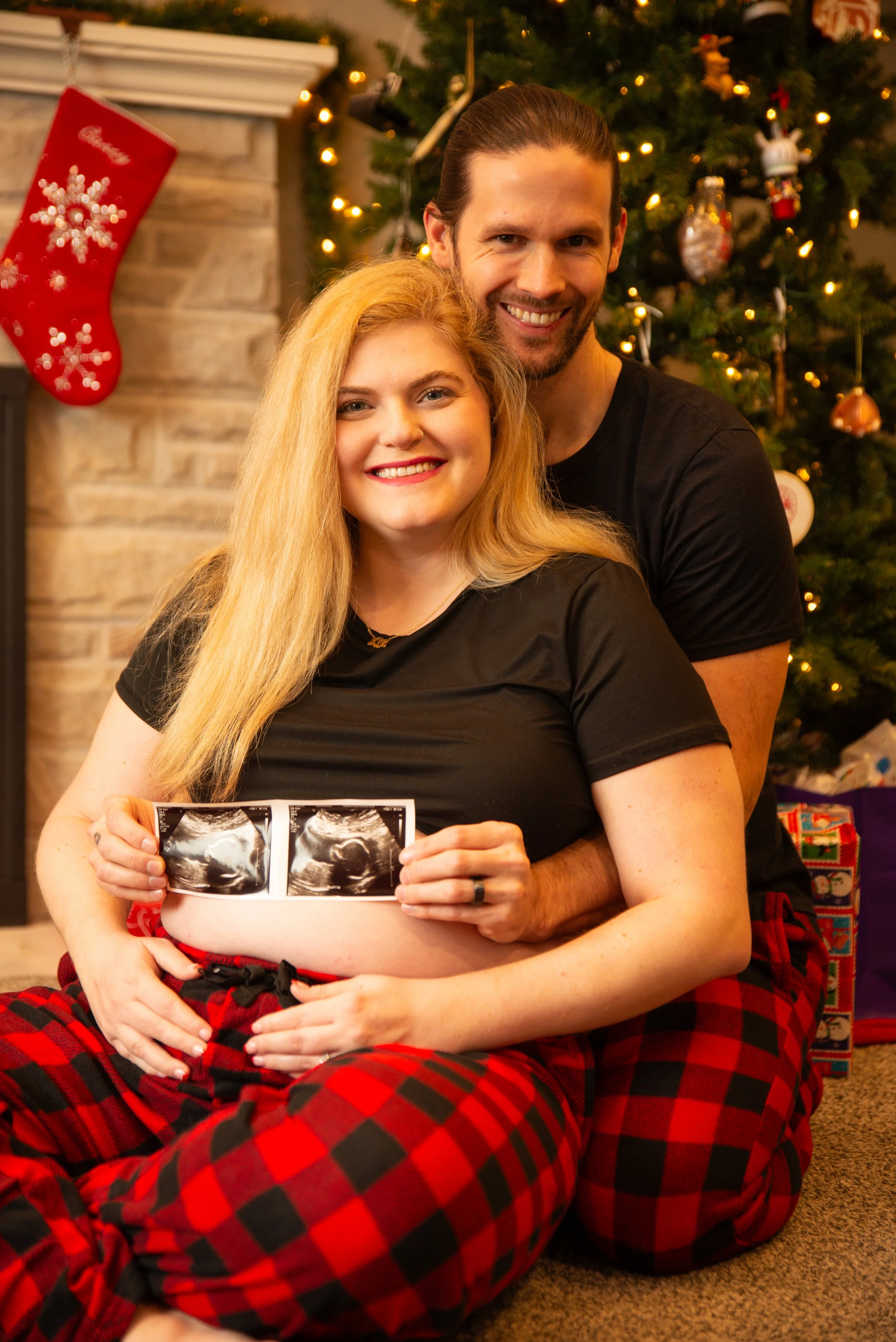 A smiling pregnant woman holding ultrasound photos, sitting on her husband's lap, in front of a Christmas tree, with her partner behind her smiling, as they celebrate Christmas.