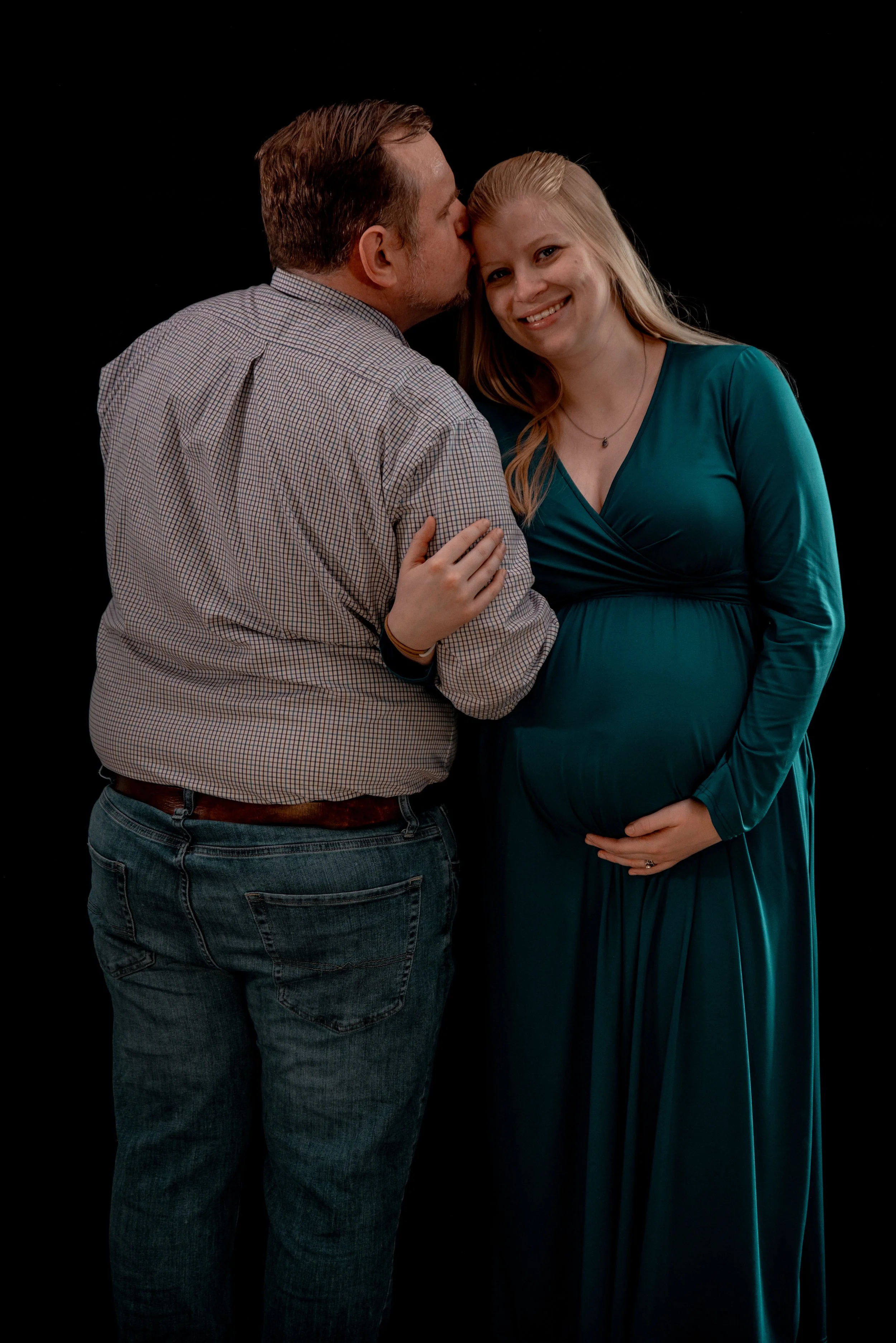 A pregnant woman in a teal dress smiles as her partner kisses her on the forehead. She is holding her belly, and they are standing against a black background.