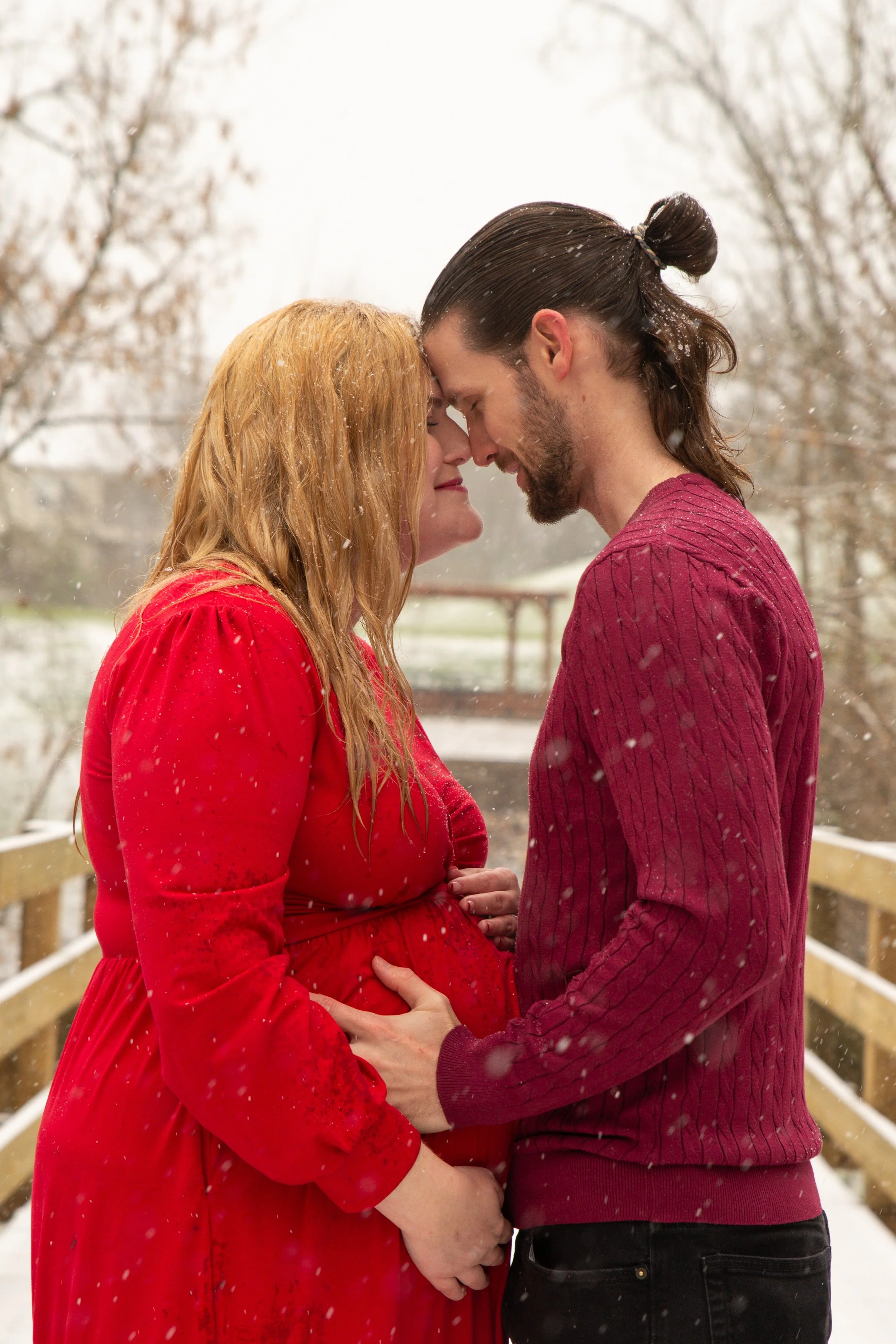 A pregnant couple with their foreheads touching, standing close on a snow-covered bridge during snowfall, with trees in the background.