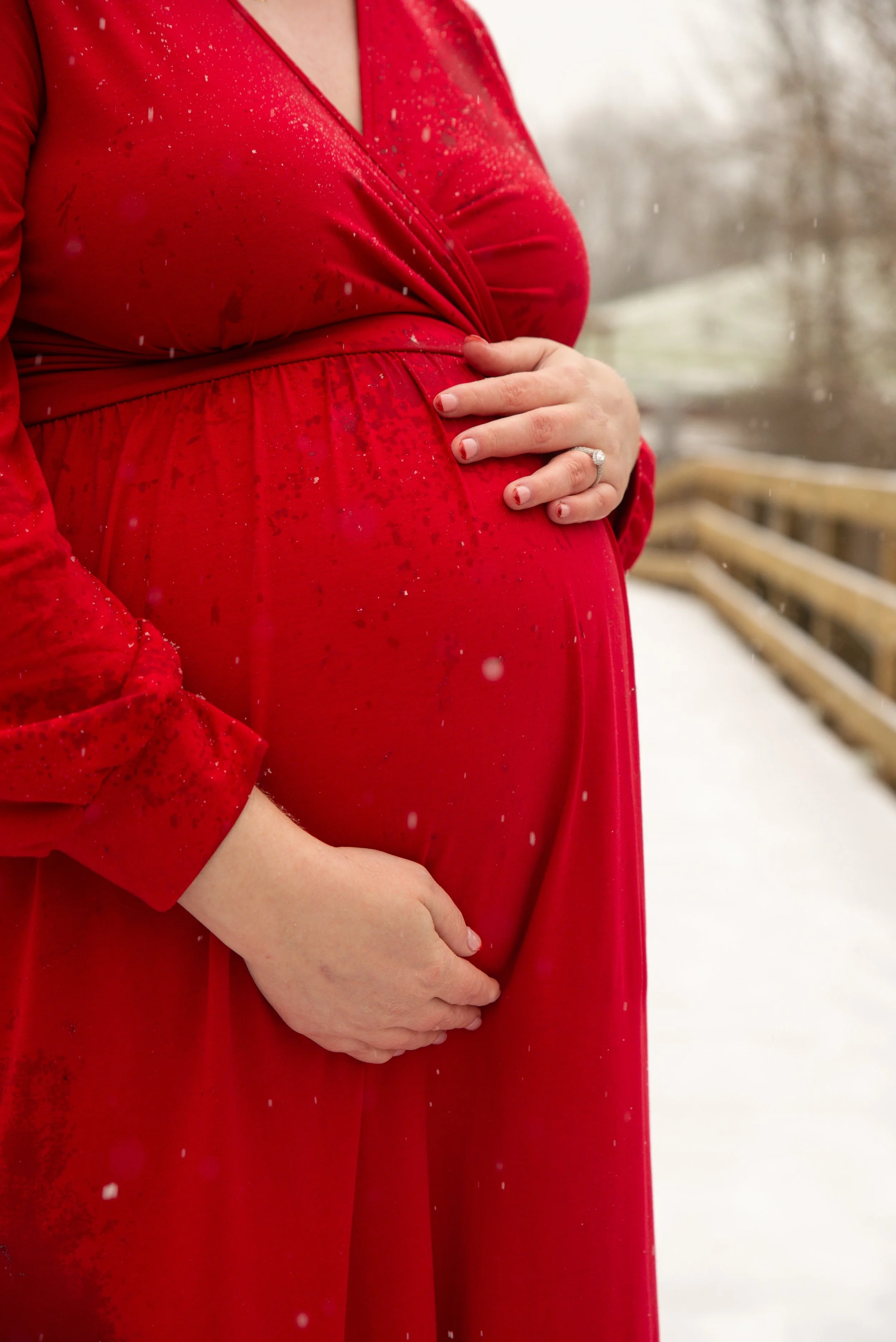 Close-up of a pregnant woman in a red dress with her hands resting on her belly, outdoors in a snowy setting.