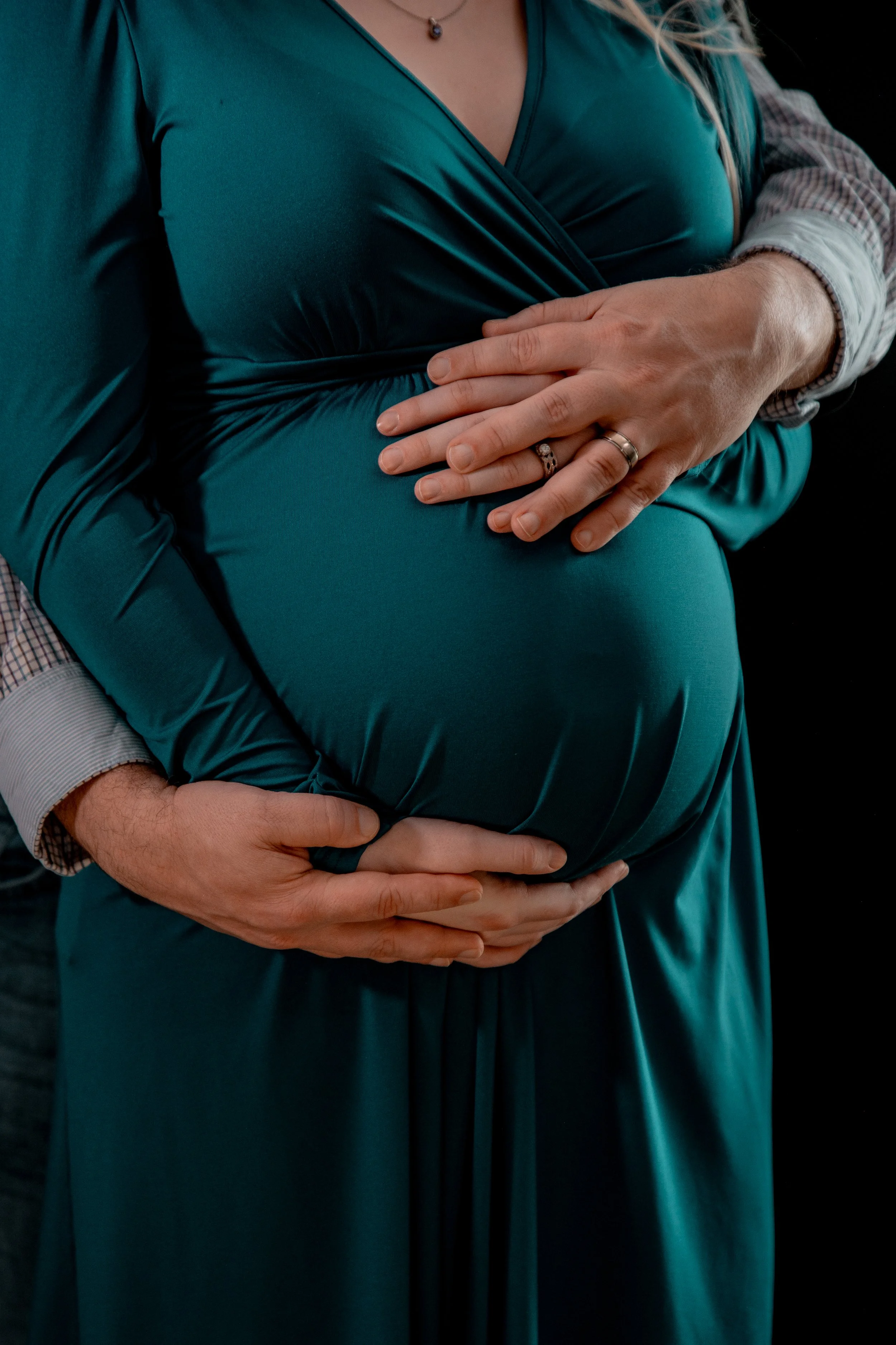 Close-up of a pregnant woman in a teal dress with her hands resting on her belly, with a man’s hands around her waist.