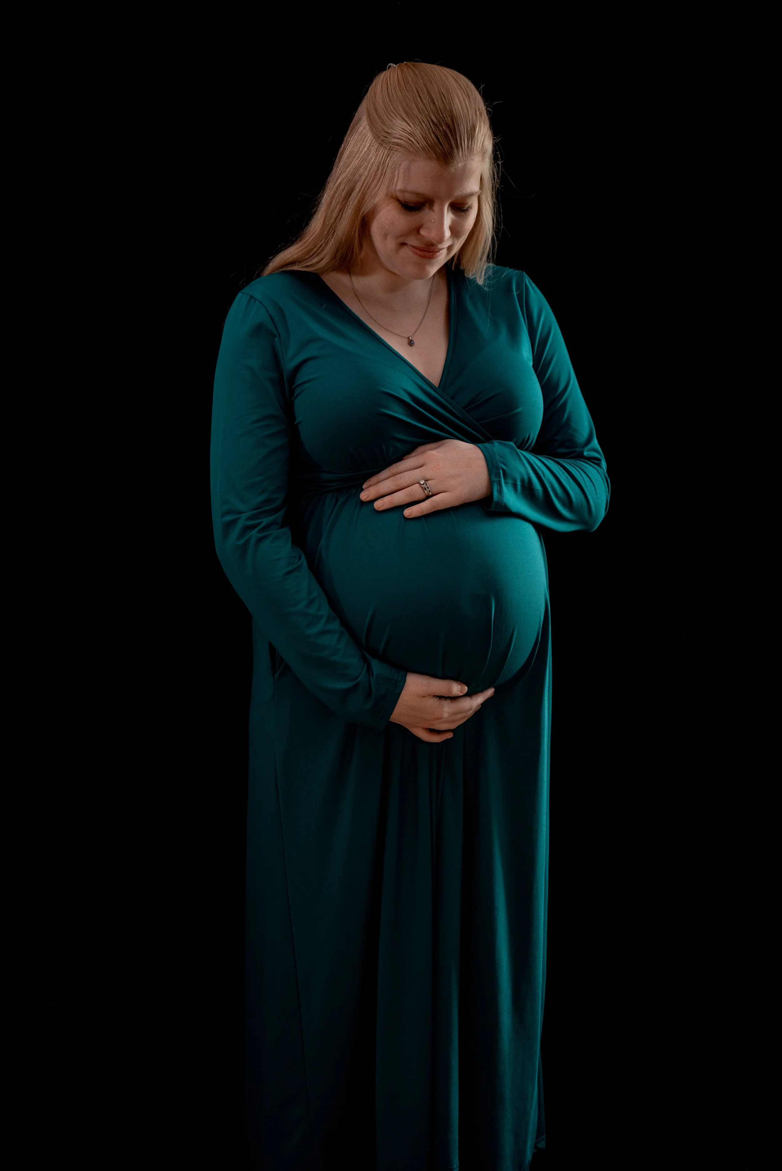 Pregnant woman in teal dress cradling her belly against a black background.