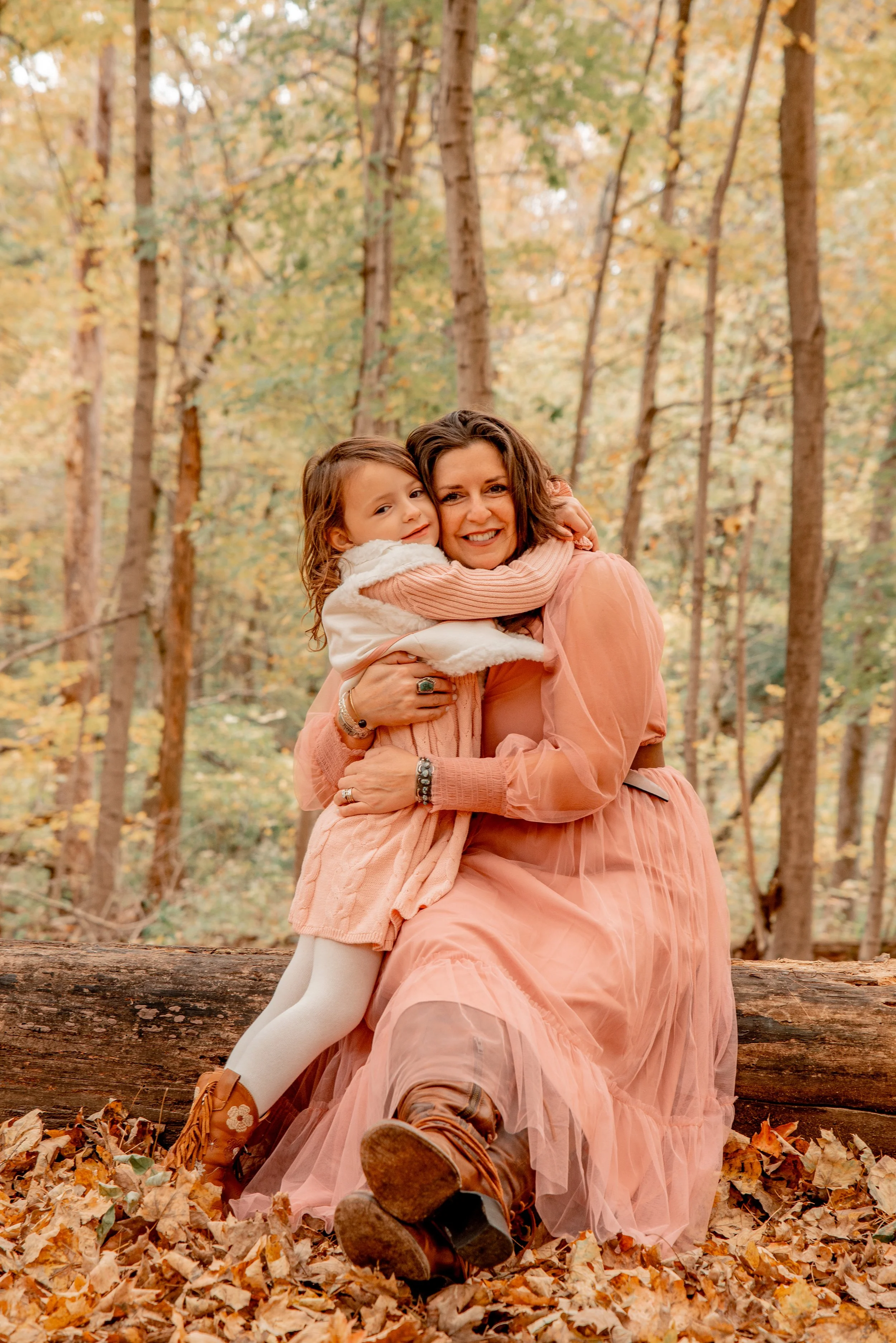 A woman and young girl sitting on a fallen log in a forest during fall, smiling and hugging, surrounded by autumn leaves and trees with colorful foliage.