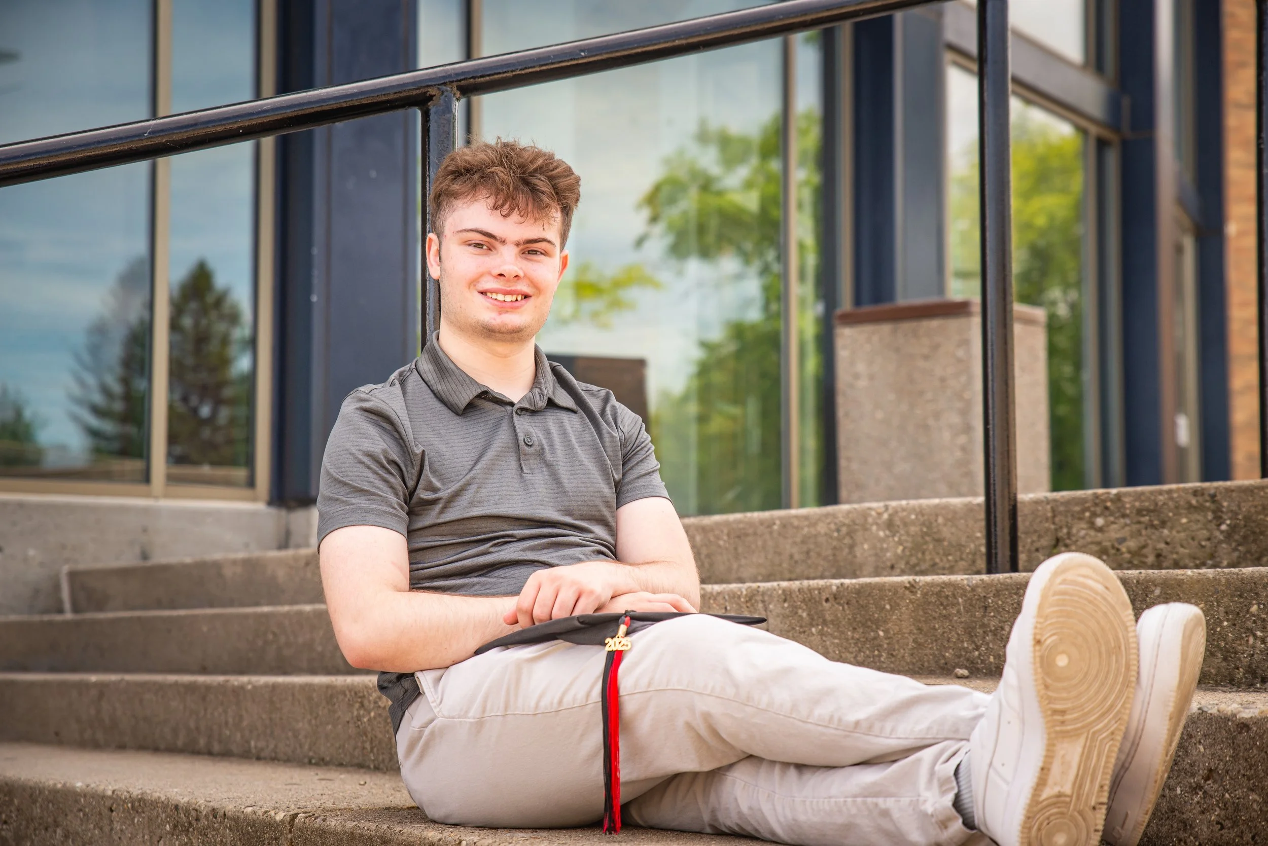 A young man sitting on outdoor steps near a building, smiling and holding a black folder with a red ribbon and a gold charm attached, wearing a gray polo shirt and beige pants.