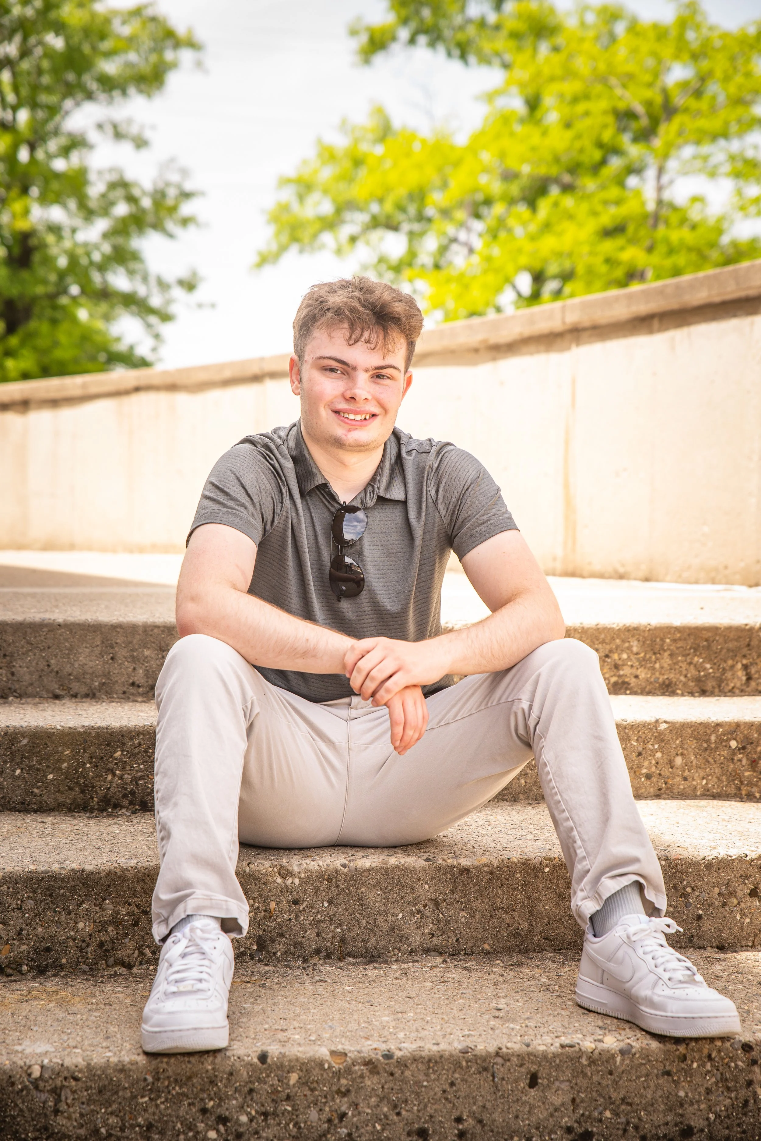 A young man sitting on outdoor concrete stairs, smiling at the camera, wearing a gray polo shirt, light-colored pants, and white sneakers, with sunglasses hanging from his shirt, under a sunny sky with green trees in the background.