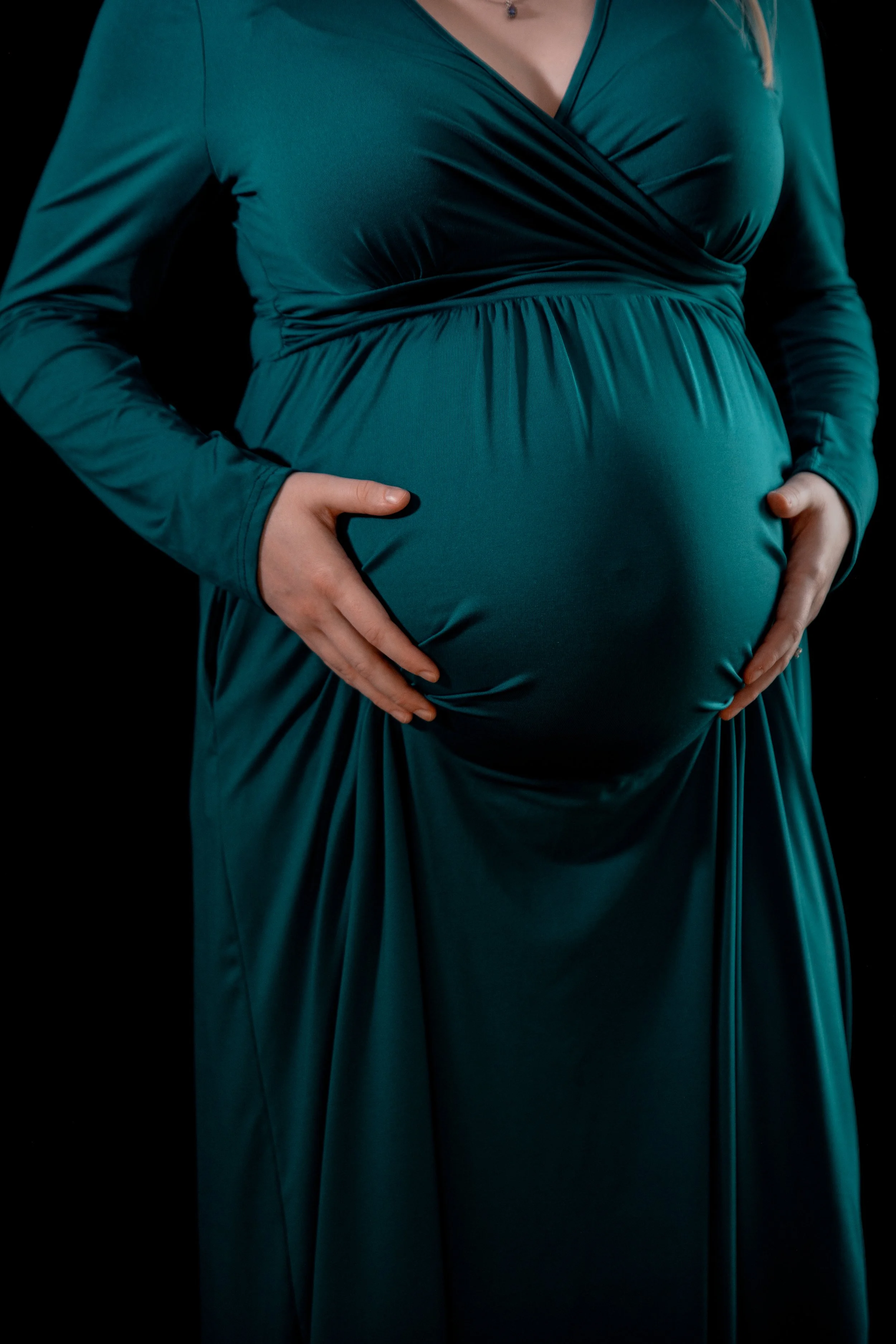 A pregnant woman wearing a teal dress with long sleeves, holding her belly with both hands against a black background.