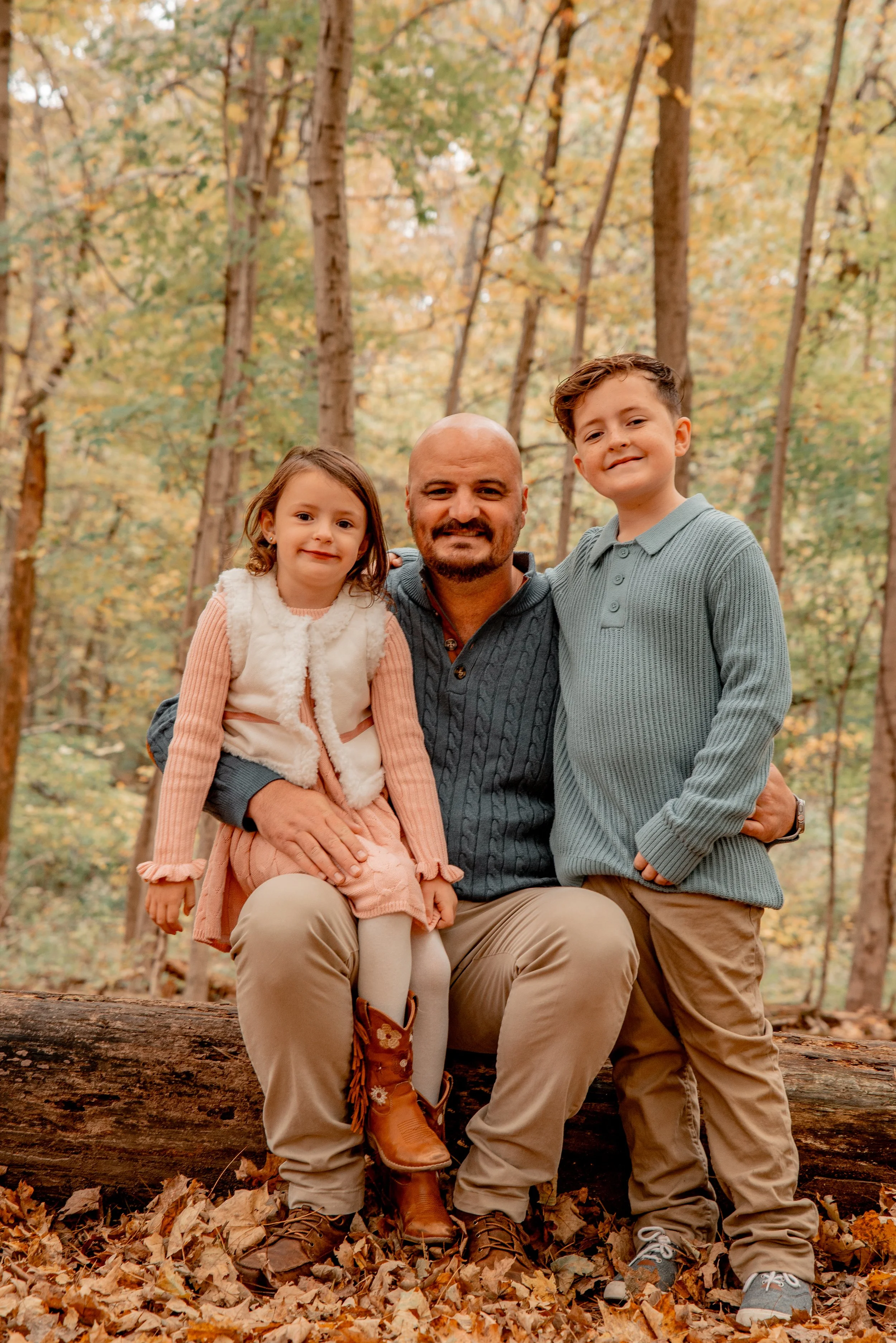 A man sitting on a fallen log in a forest during autumn, holding a young girl on his lap and standing next to a boy, all smiling at the camera. The forest has tall trees with yellow and orange leaves.