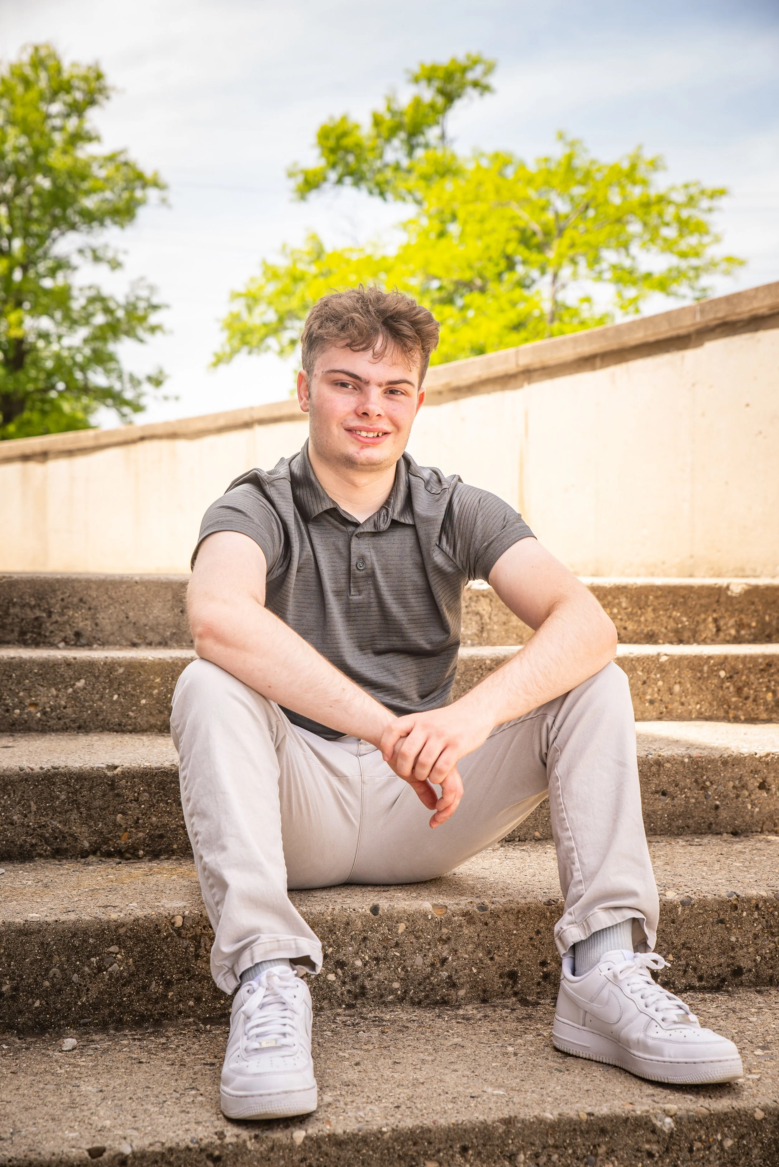 A young man sitting on outdoor concrete steps, smiling, wearing a dark gray polo shirt, light gray pants, and white sneakers, with green trees and a cloudy sky in the background.