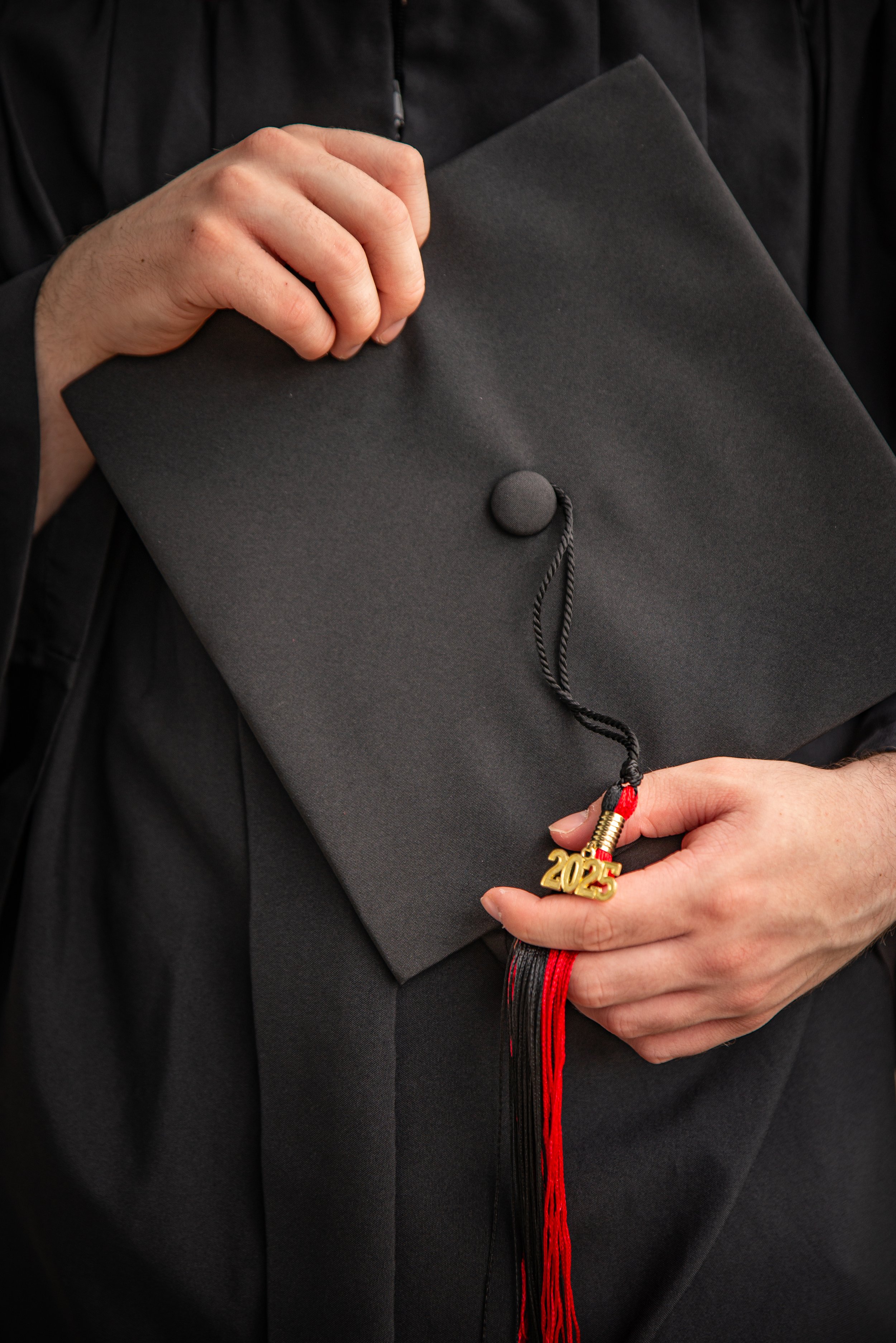A person holding a black graduation cap with a tassel that has a gold "2025" charm, wearing a black graduation gown.