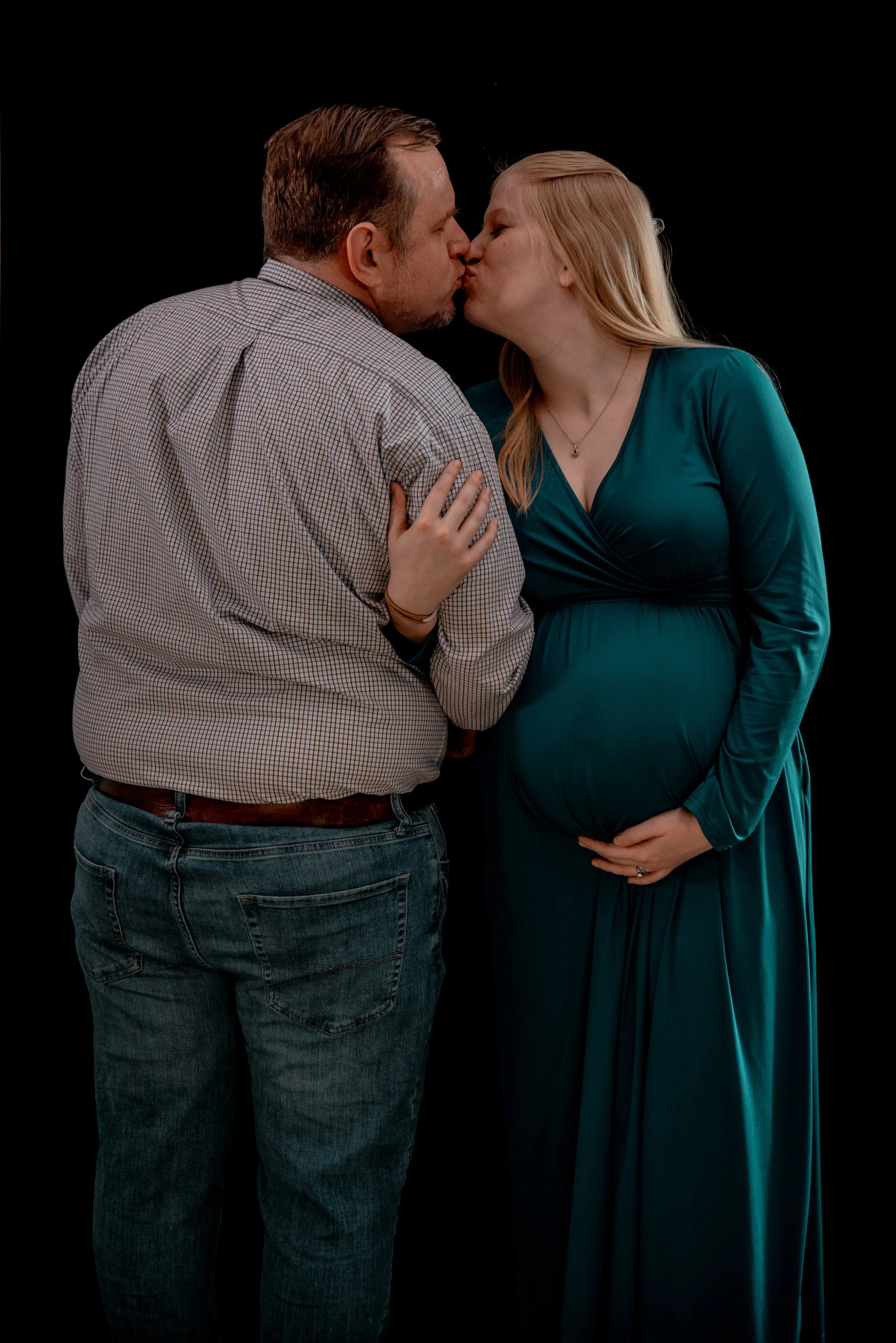 A pregnant woman in a teal dress and a man with a checkered shirt kiss against a black background.