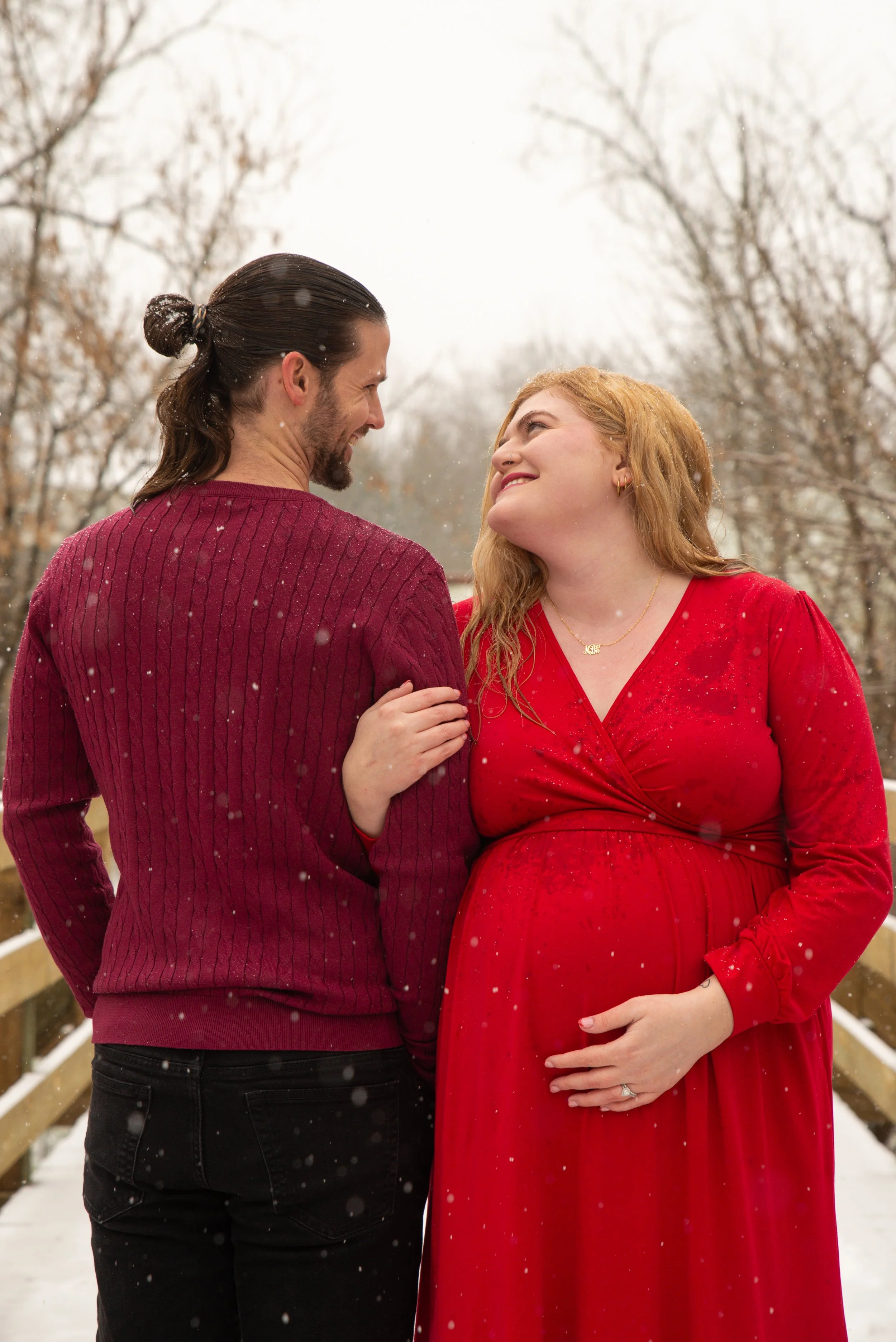 A couple standing close together outdoors in winter, smiling at each other, with snow falling around them. The man has long dark hair tied in a bun, wearing a red sweater, and the woman has red hair, wearing a red dress and a gold necklace.