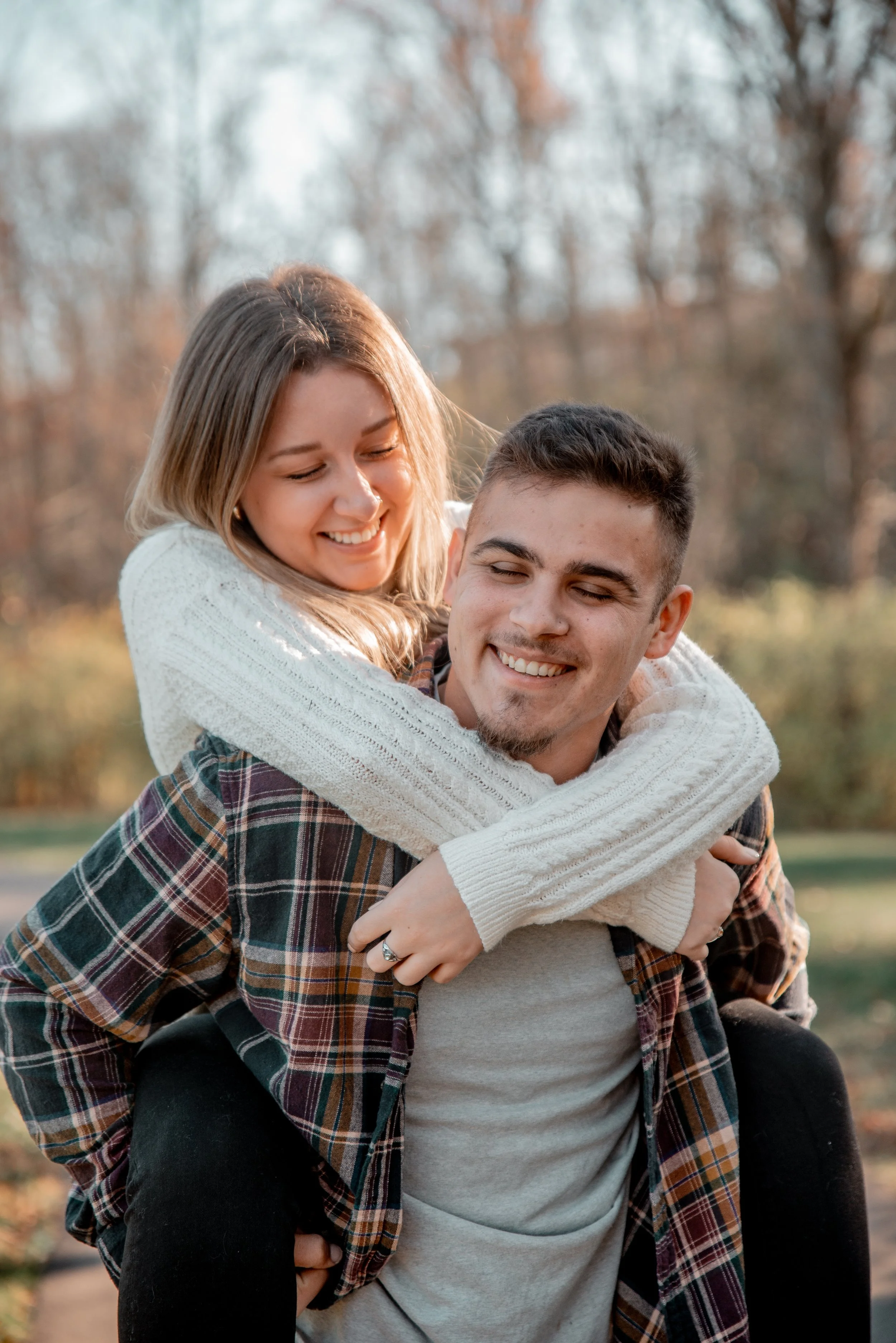 A young woman with blonde hair and a white sweater is riding on the back of a young man with dark hair, a beard, and a plaid shirt. They are outdoors in a park with autumn trees in the background, both smiling and enjoying each other's company.