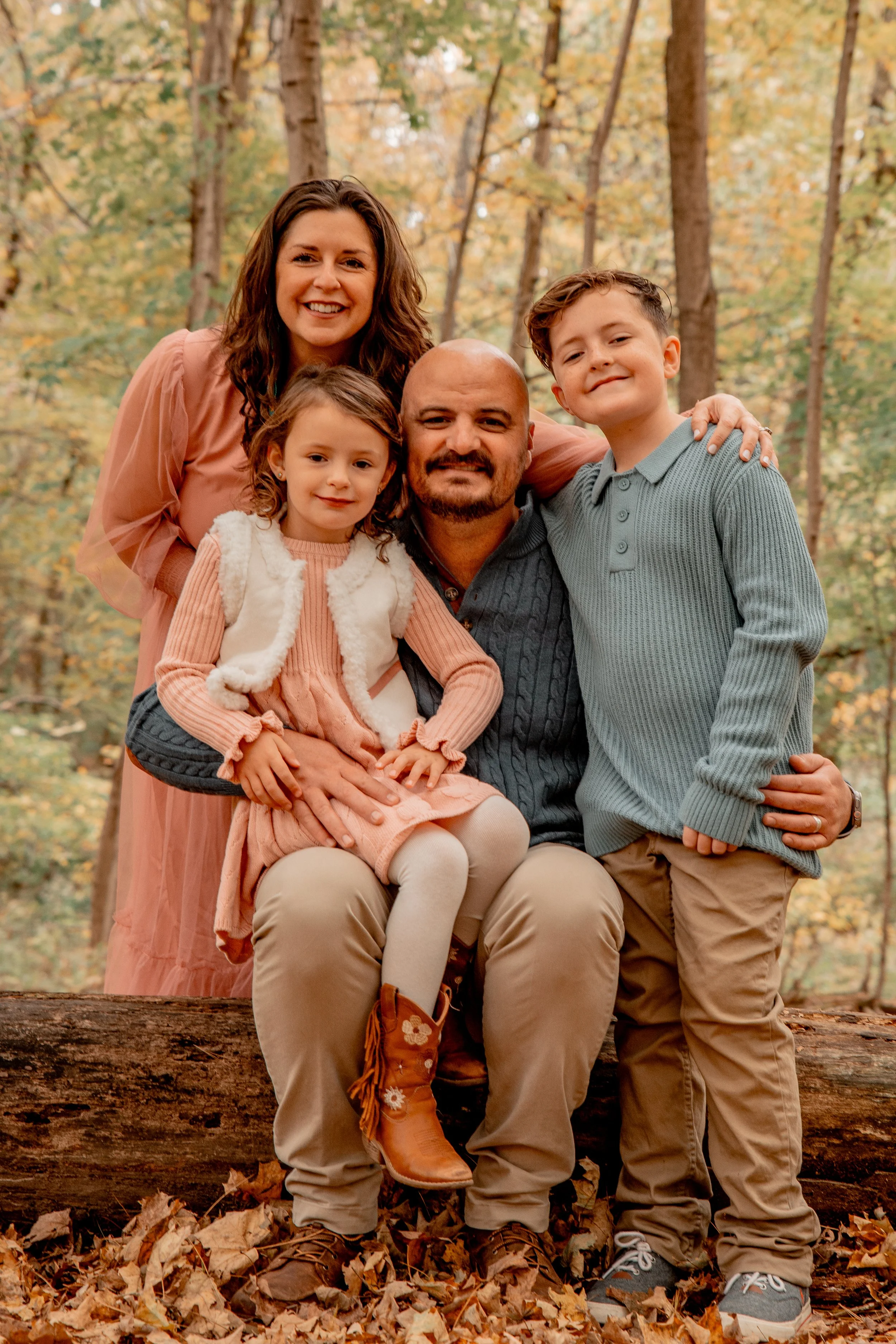 A family of five posing outdoors in an autumn forest, smiling. The mother with curly hair stands behind, the father has a beard and is seated on a log, holding a young girl on his lap. The girl wears a pink dress and boots. The son, wearing a light b