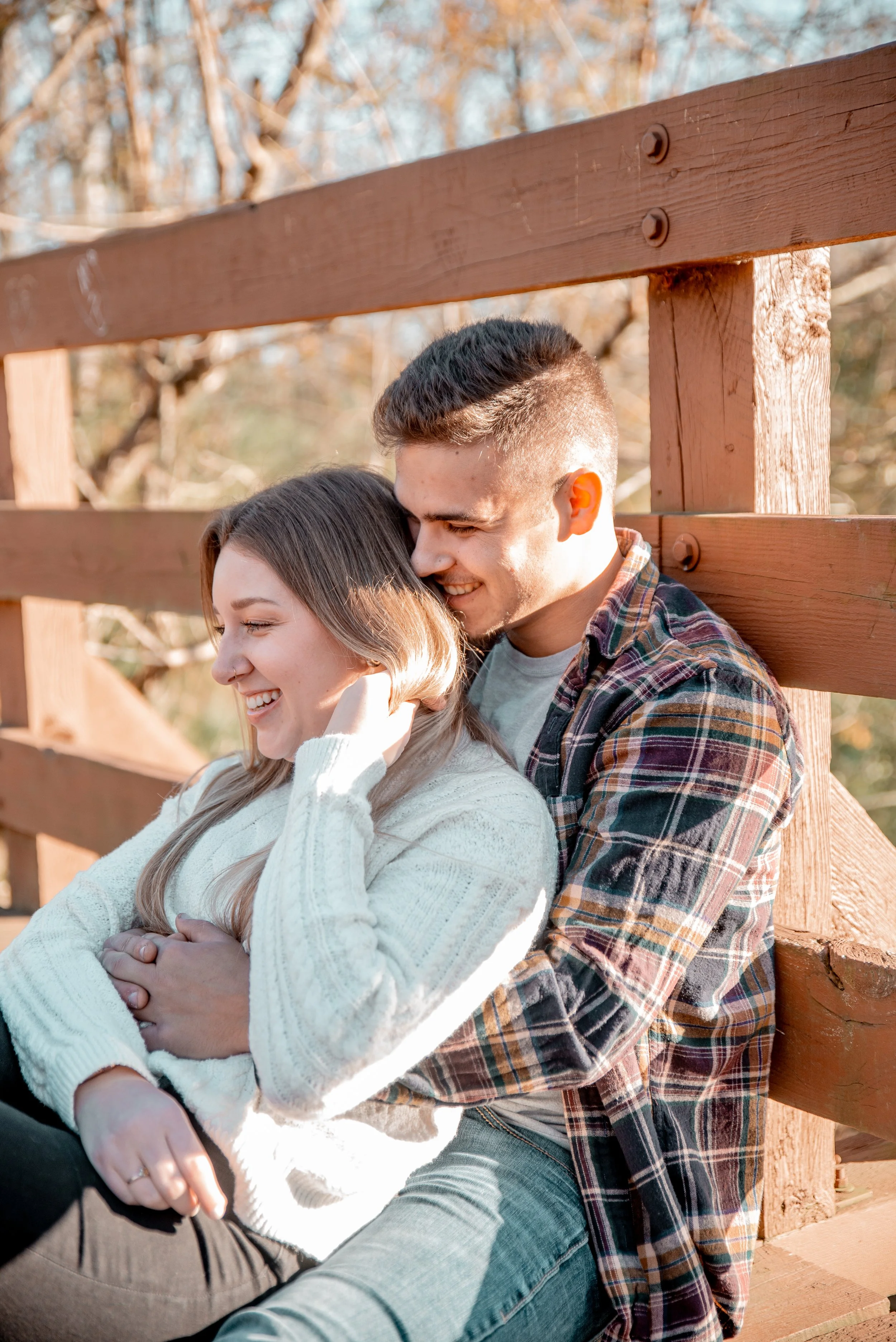 A smiling young couple sitting outdoors against a wooden fence, enjoying a sunny day.