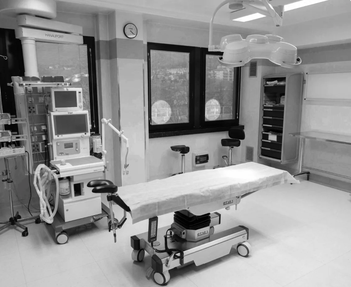 Stanza di un ospedale con letto operatorio, monitor, lampada chirurgica, armadietto e finestra.