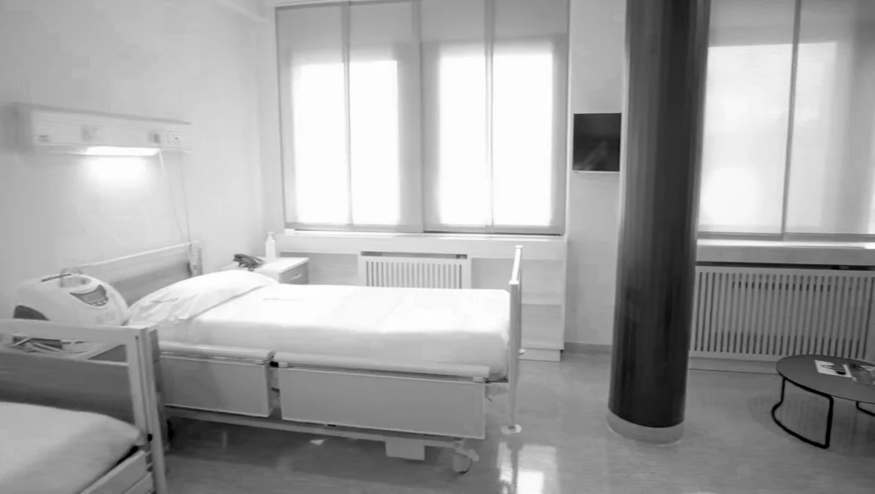 Stanza di ospedale con due letti, una finestra grande, un tavolo rotondo e uno scaffale.