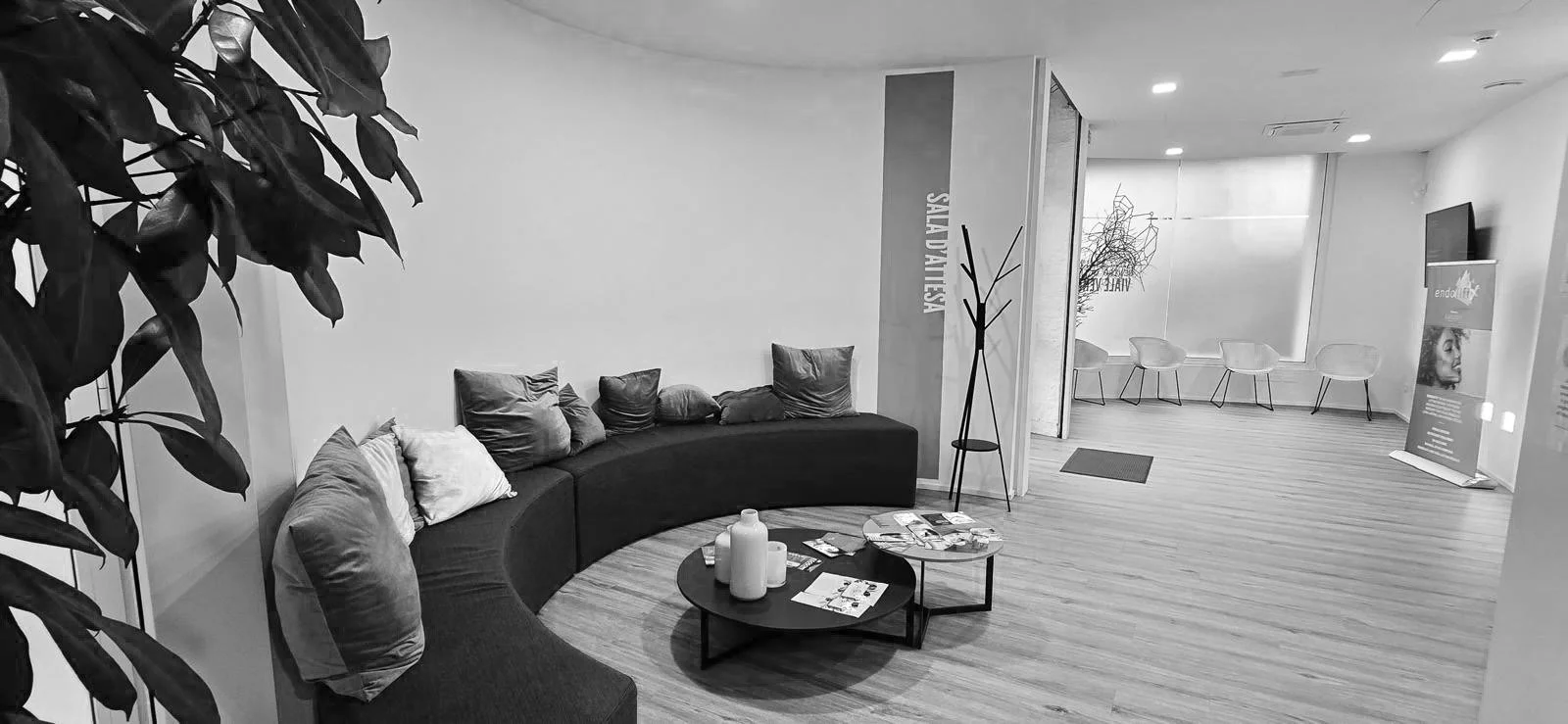 Sala d'attesa con divano curvo, cuscini, tavolini con riviste e brocche, sedie lungo la finestra, panca con cartelli informativi, e decorazioni minimaliste.