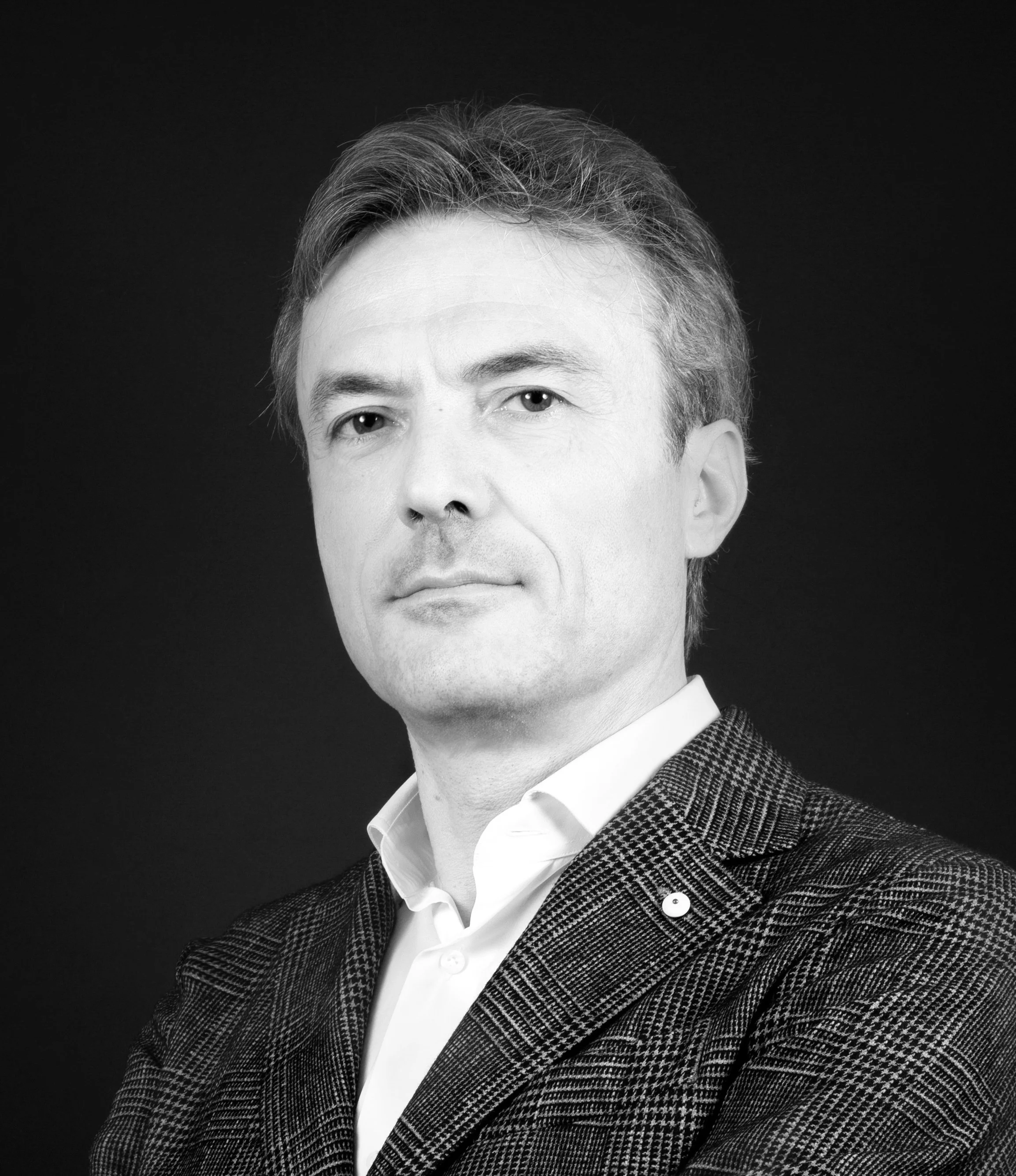 Ritratto in bianco e nero di un uomo con capelli castani chiari, che indossa una camicia bianca e una giacca con motivo check, con sfondo scuro.