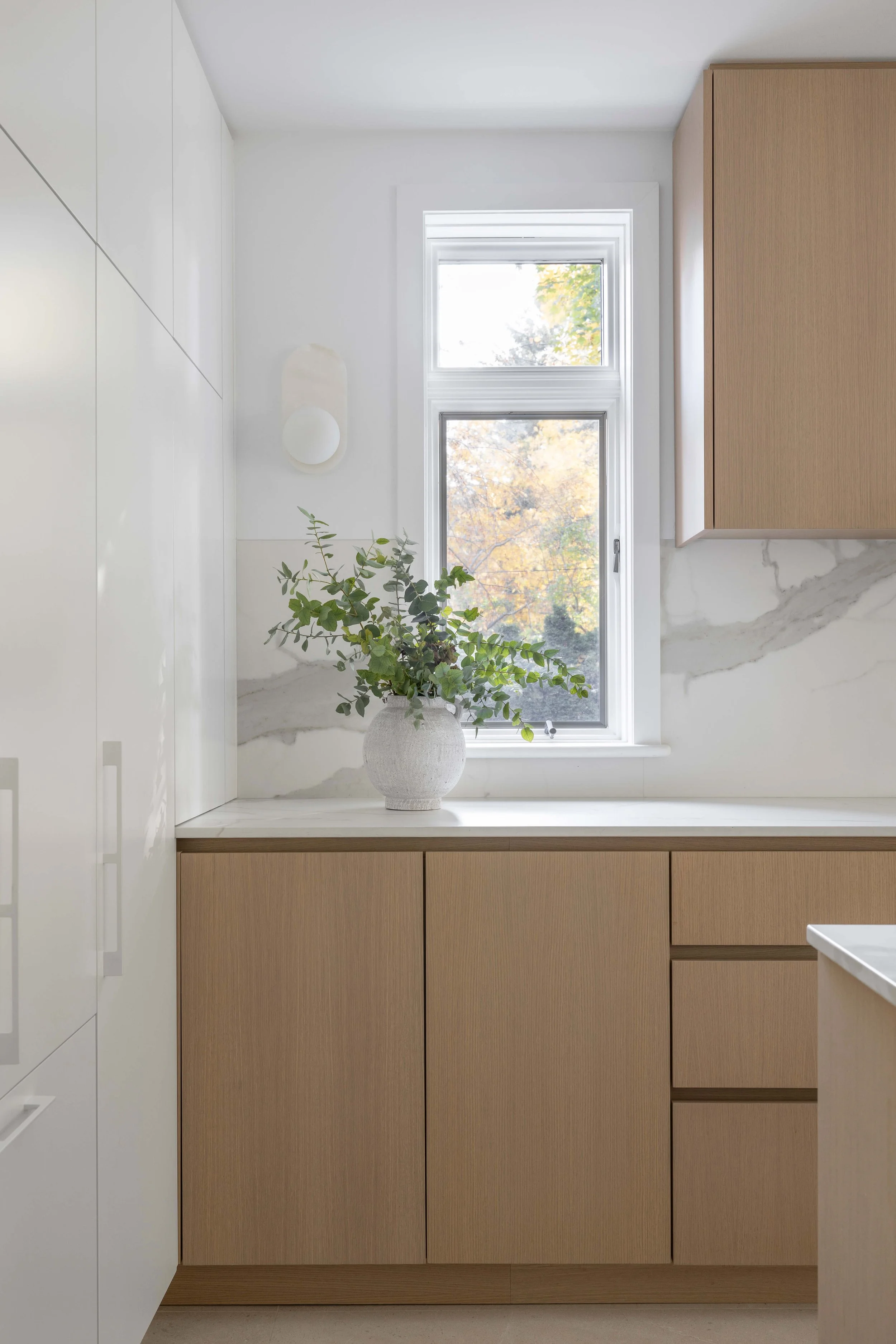 Une cuisine moderne avec des placards en bois clair, un vase blanc avec des plantes vertes sur une surface en marbre sous une fenêtre laissant entrer la lumière naturelle.