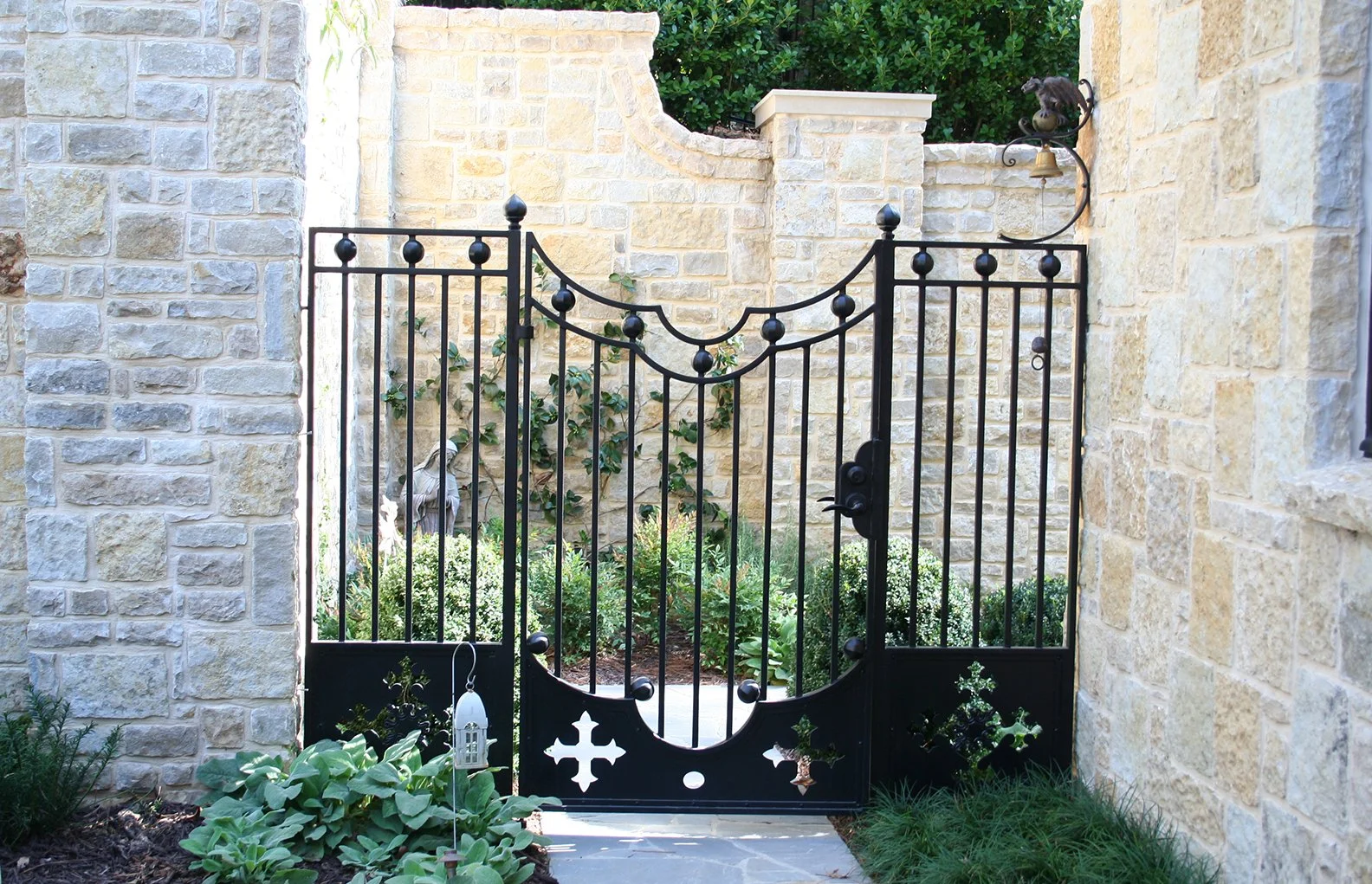A black wrought iron gate with decorative spheres and cross cutouts, set between stone walls, leading into a garden with plants and a bird statue.