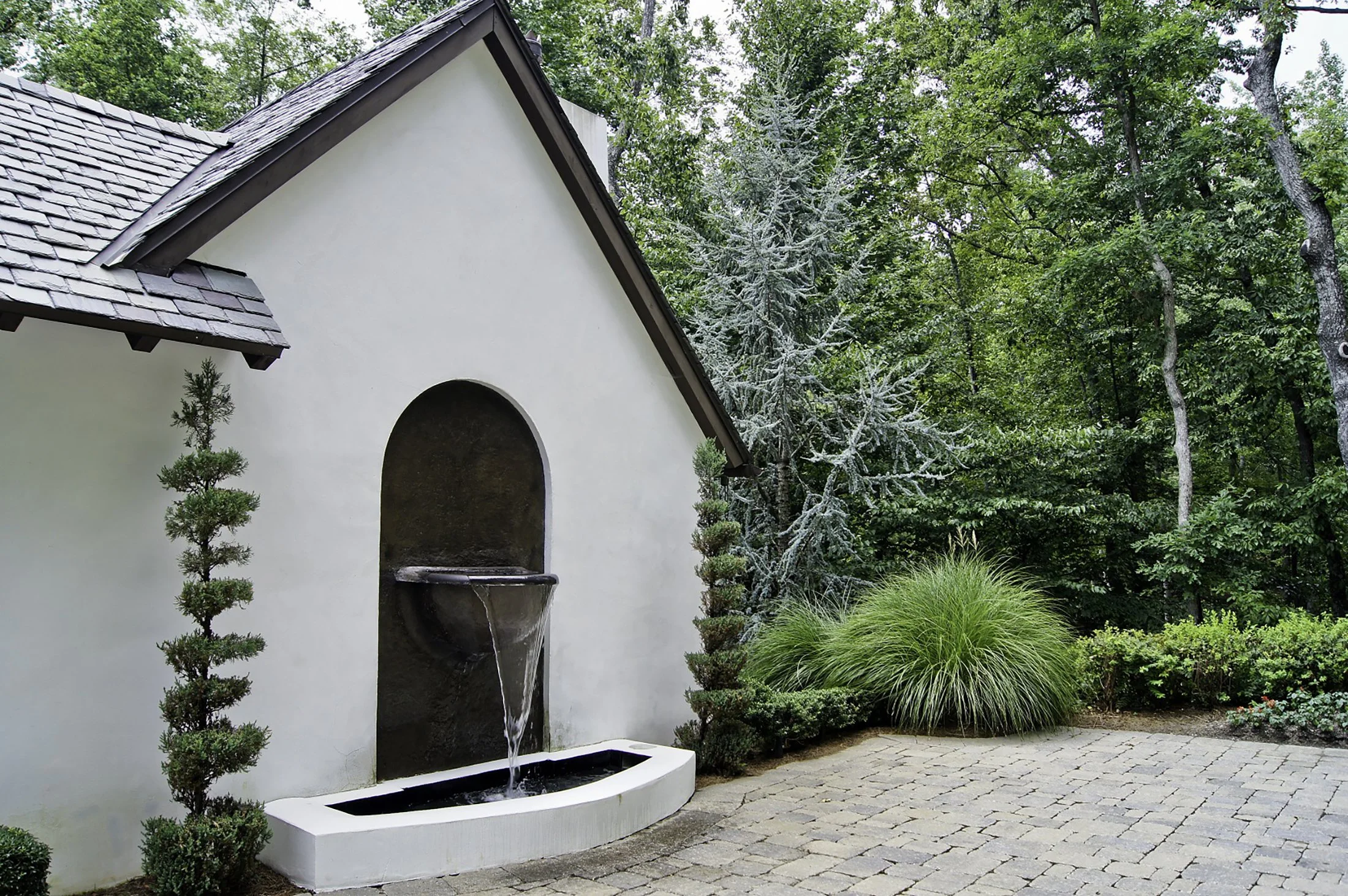 White outdoor wall with a small fountain, surrounded by green plants and trees in a garden setting.