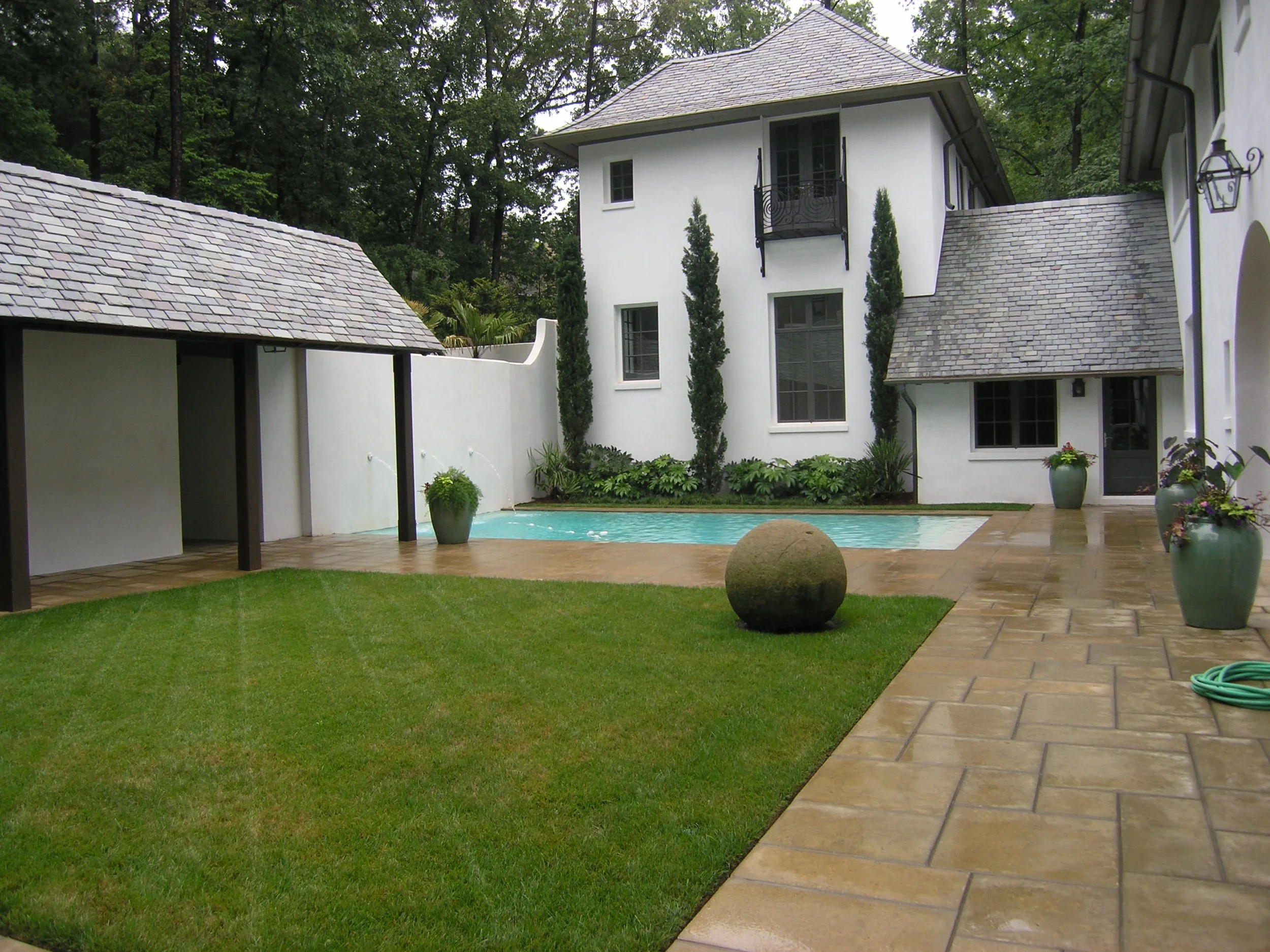 Residential backyard with a small swimming pool, grassy area, potted plants, stone pathway, and a white house with gray shingle roof and black balcony.