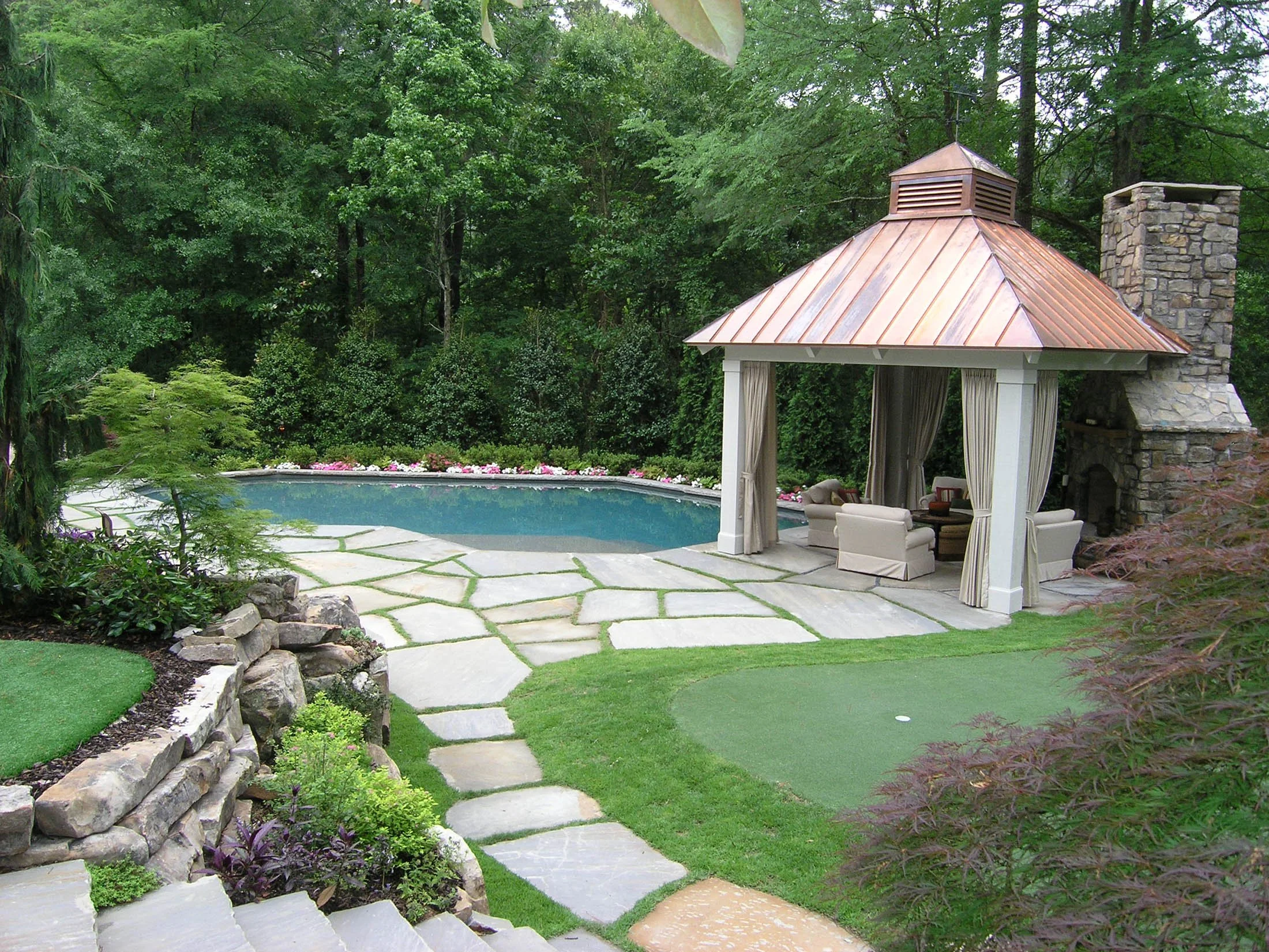 A backyard with a swimming pool, a stone patio, lush greenery, and a gazebo with curtains and a copper roof, built near a stone chimney.