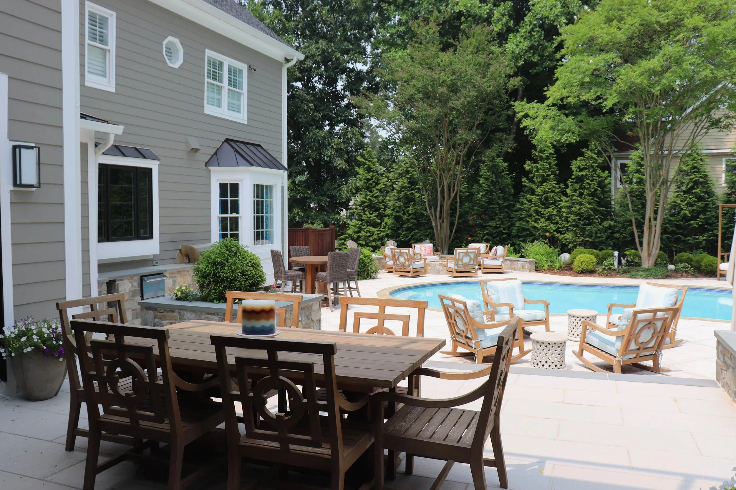 A backyard with a swimming pool surrounded by outdoor seating, including armchairs, lounge chairs, and a dining table with chairs, featuring trees and greenery in the background.