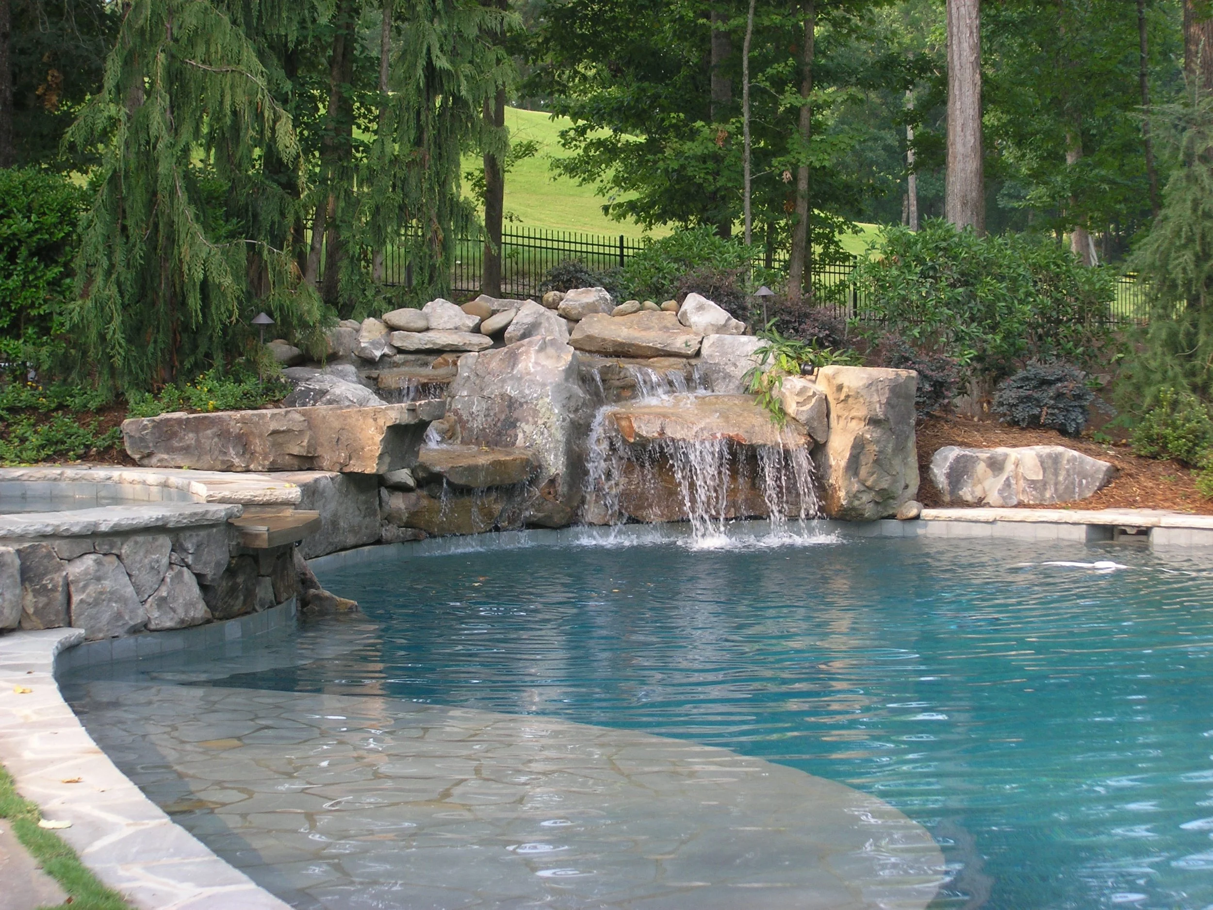 A backyard with a swimming pool and a water feature made of rocks, small waterfalls, surrounded by greenery and trees.