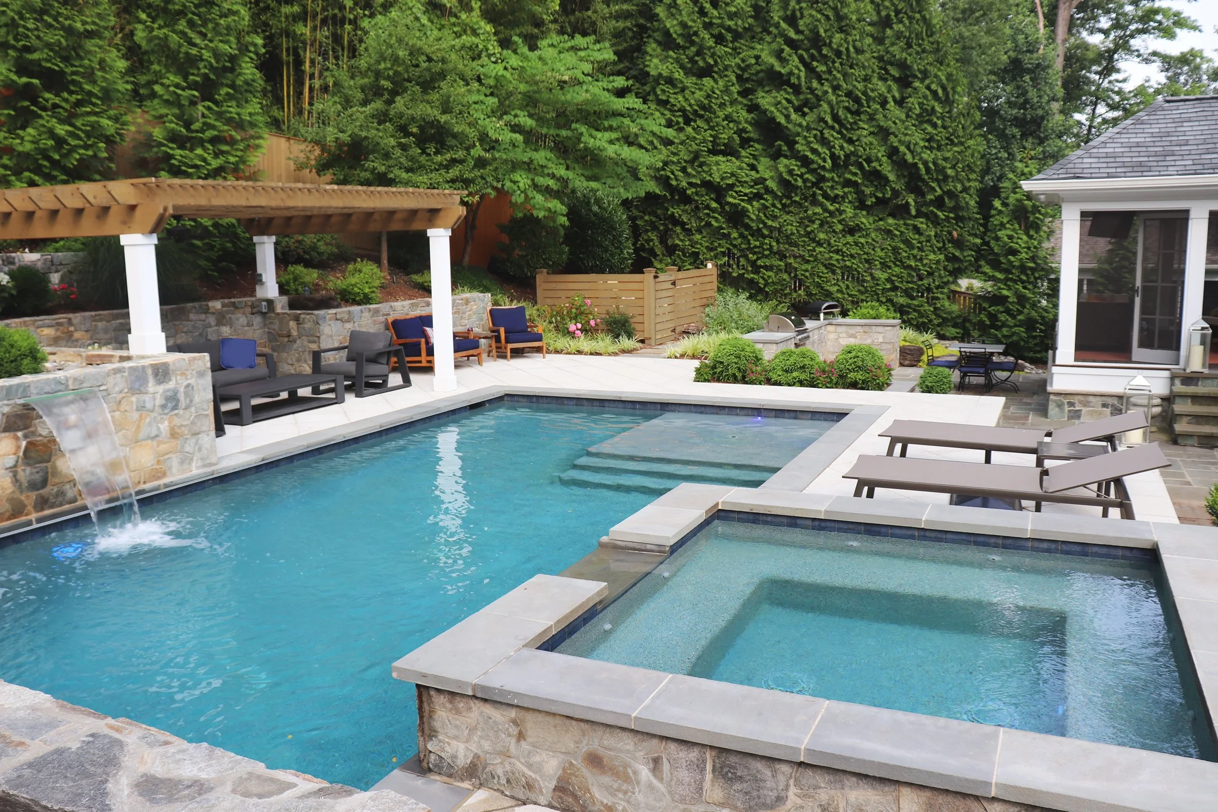 A backyard with a swimming pool, hot tub, relax chairs, a shaded seating area, lush greenery, and a house in the background.